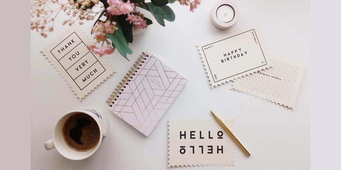 Flat lay of a pink and white notepad, gold pen, and three handwritten notecards reading "Thank You Very Much", "Happy Birthday", and "Hello", surrounded by a coffee cup, flowers, and a tea light candle on a white desk.