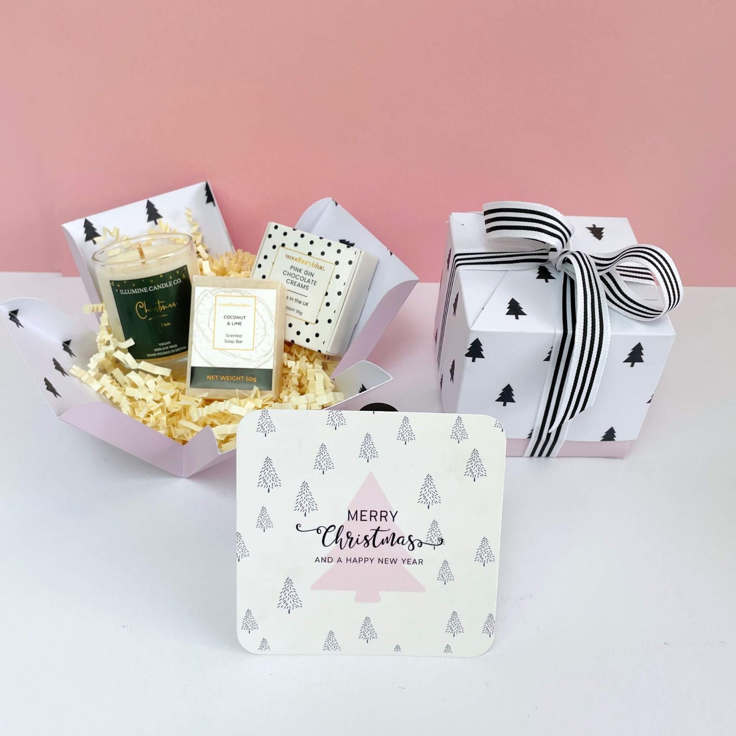 Open and closed Warm Wishes Gift Box with shredded filling, festive notecard, candle, soap, and chocolates, styled on pink backdrop.