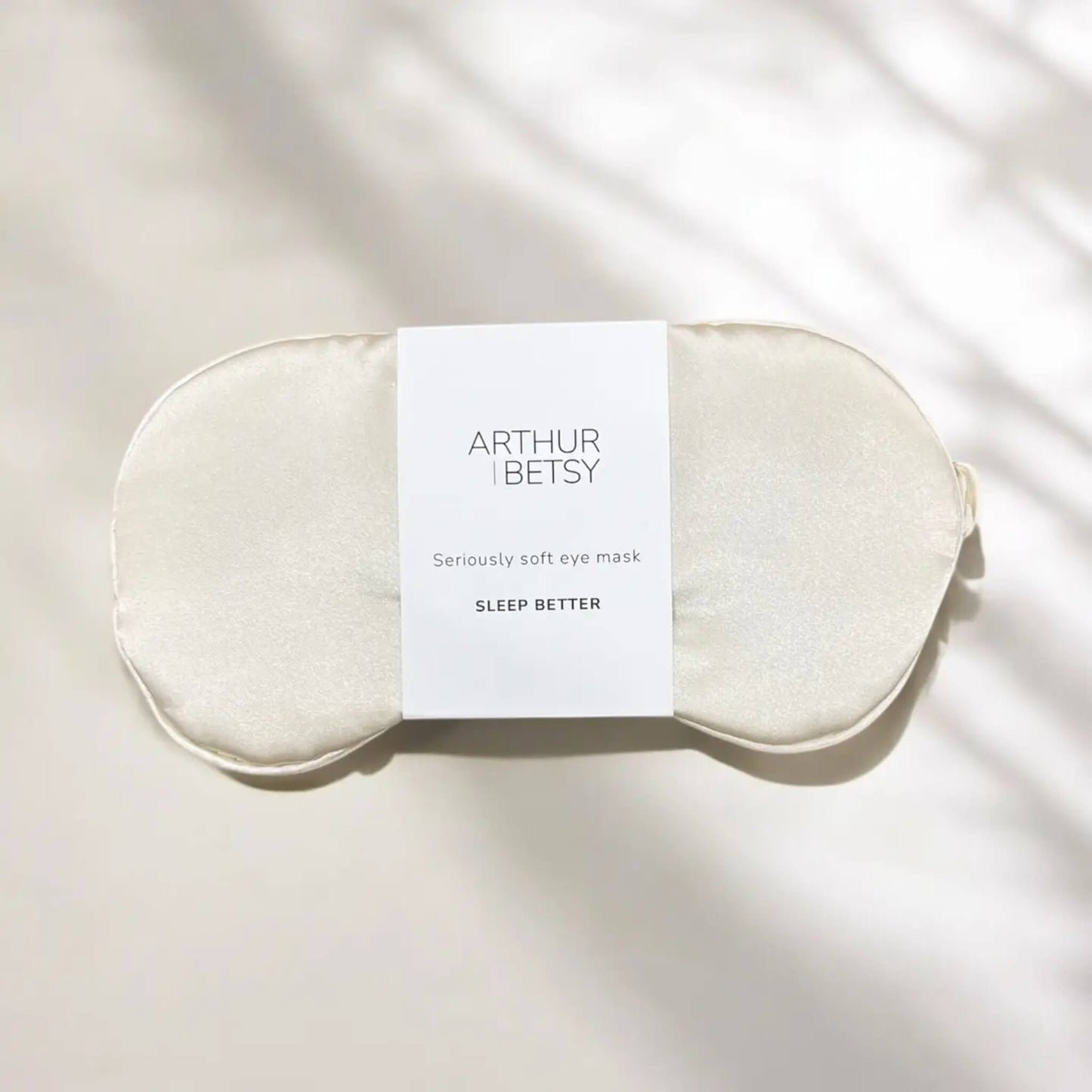 Flat lay of ivory satin sleep mask by Arthur Betsy, resting on a soft cream surface in natural light