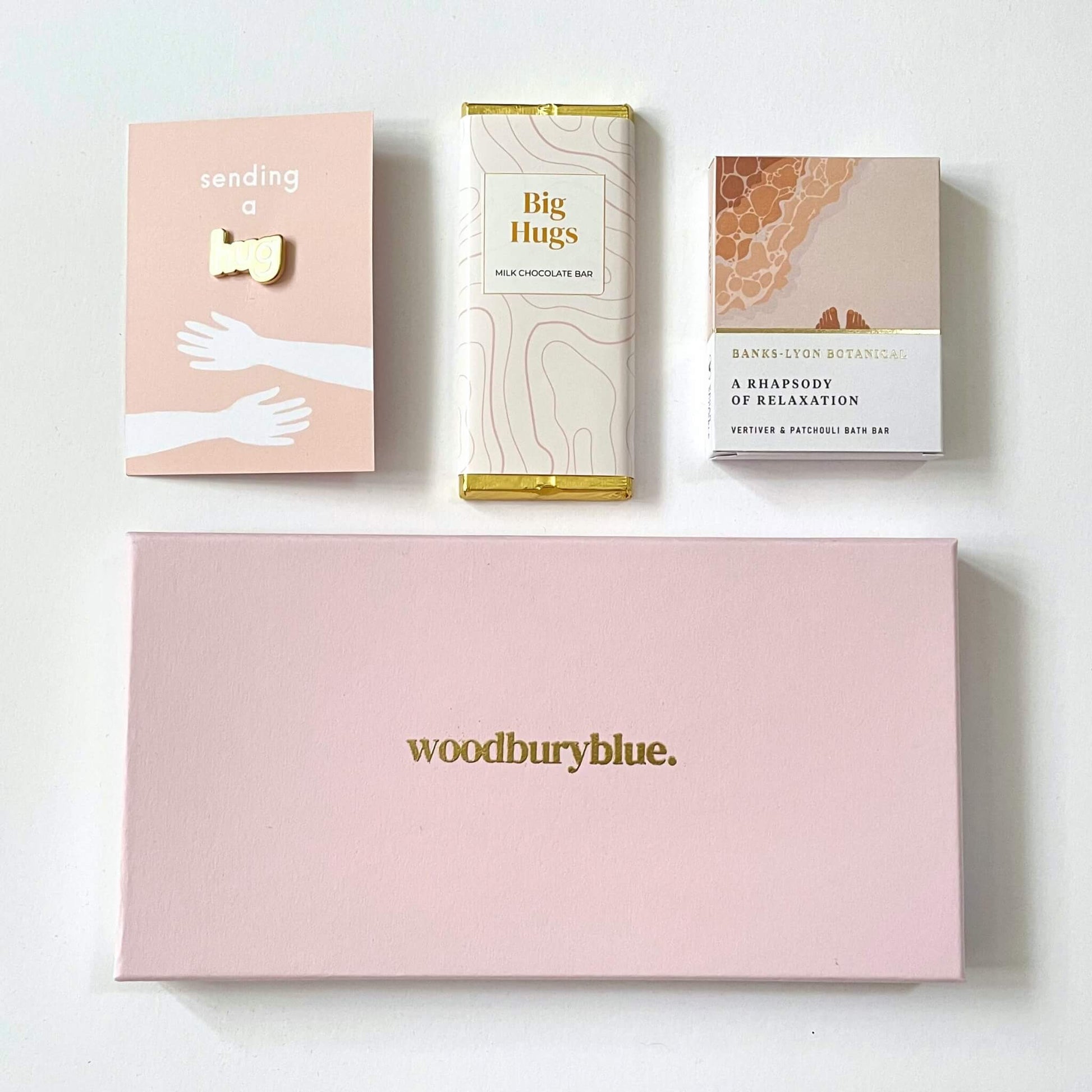 Flat lay showing A Hug in a Letterbox Gift, big hugs chocolate bar, enamil pin badge and a bath oil bar.