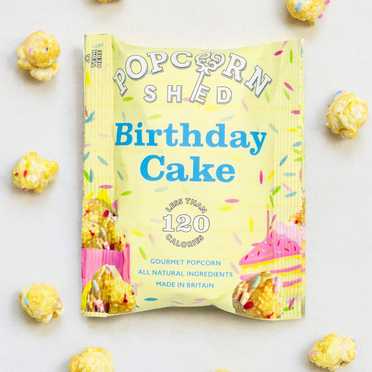 Popcorn Shed Birthday Cake Gourmet Popcorn 24g sachet on a pale background with rainbow sprinkle popcorn pieces scattered around and natural shadows.