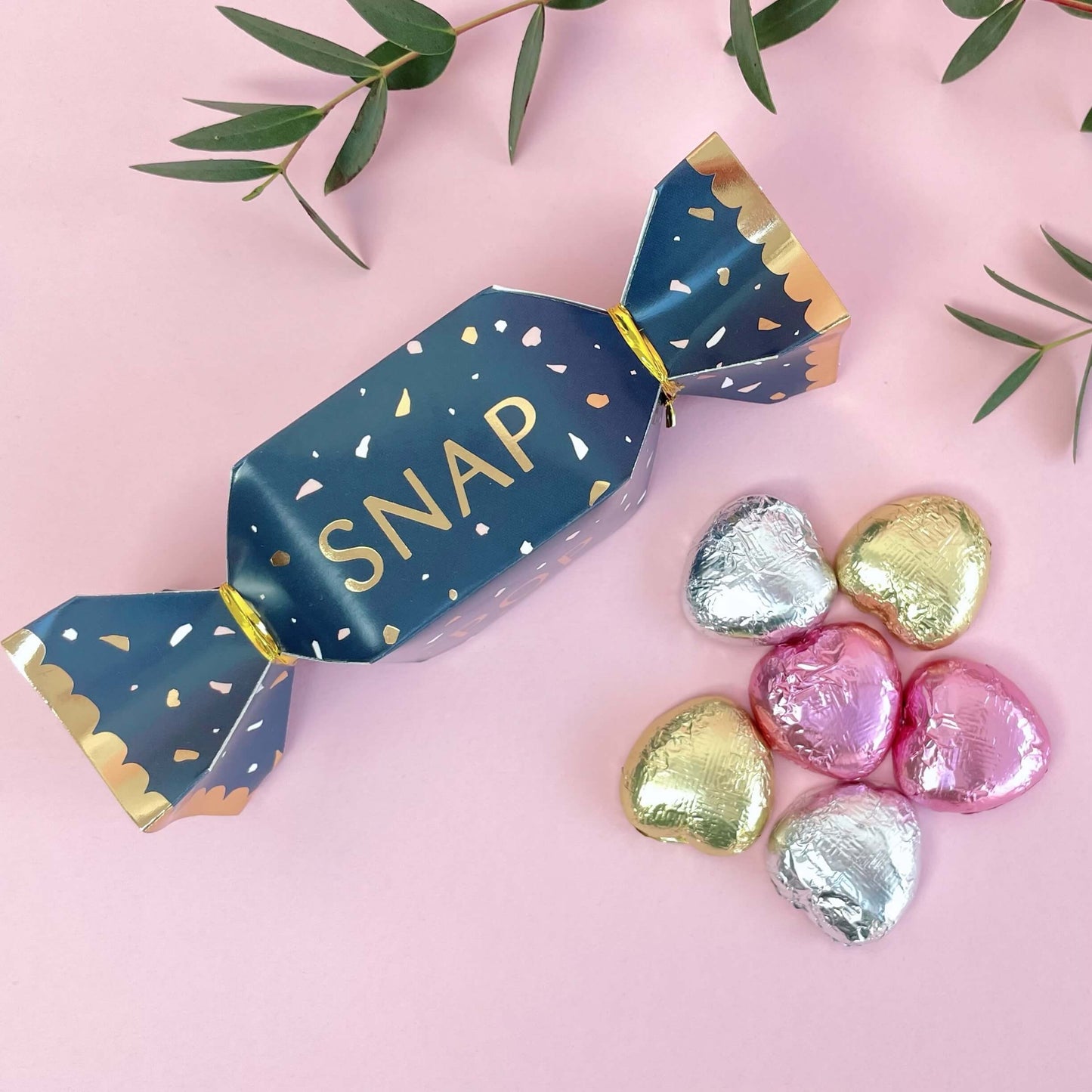 Blue and gold christmas cracker with foil wrapped milk chocolate hearts on a pink background