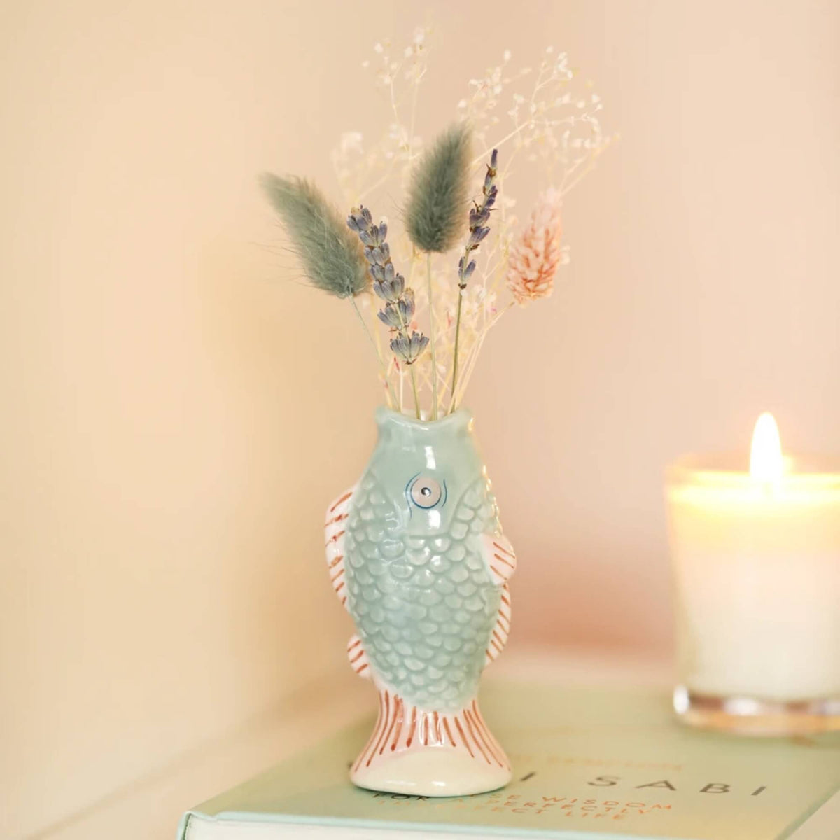 Small blue ceramic fish bud vase with dried lavender and flowers, displayed on a book beside a lit candle.