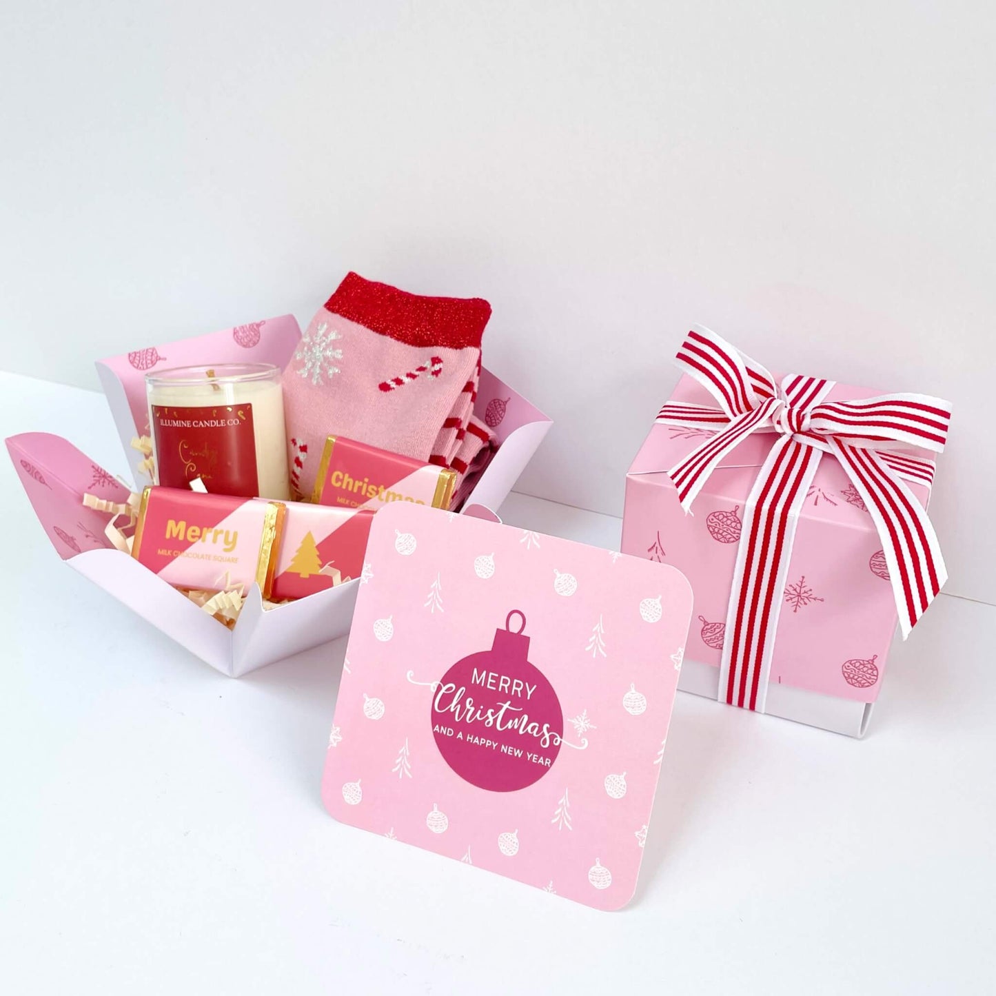 Unboxed Candy Cane Wishes Gift Box with pink festive socks, candy cane candle, Merry Christmas chocolate squares, and a pink bauble card beside the closed pink ribbon-wrapped gift box.
