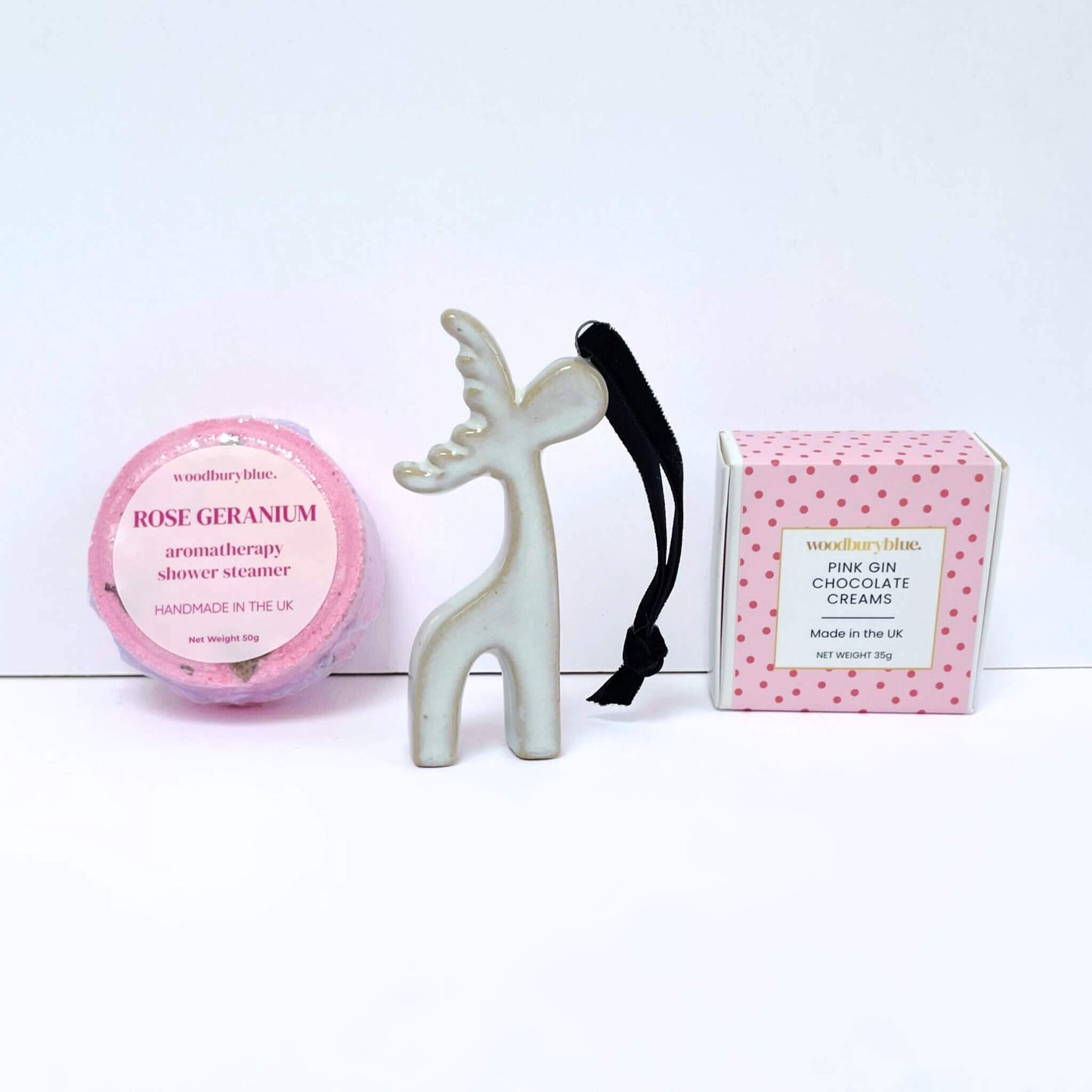 Three products on a white background: rose geranium shower steamer, a ceramic reindeer christmas tree decoration, and a pink box of chocolates.