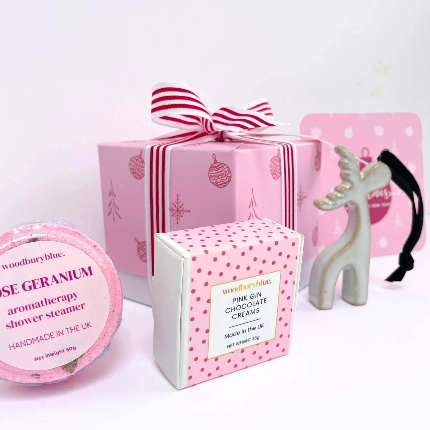 Collection of pink Christmas-themed products including a gift box with notecard, shower steamer, reindeer tree decoration and a box of chocolate creams on a white background.