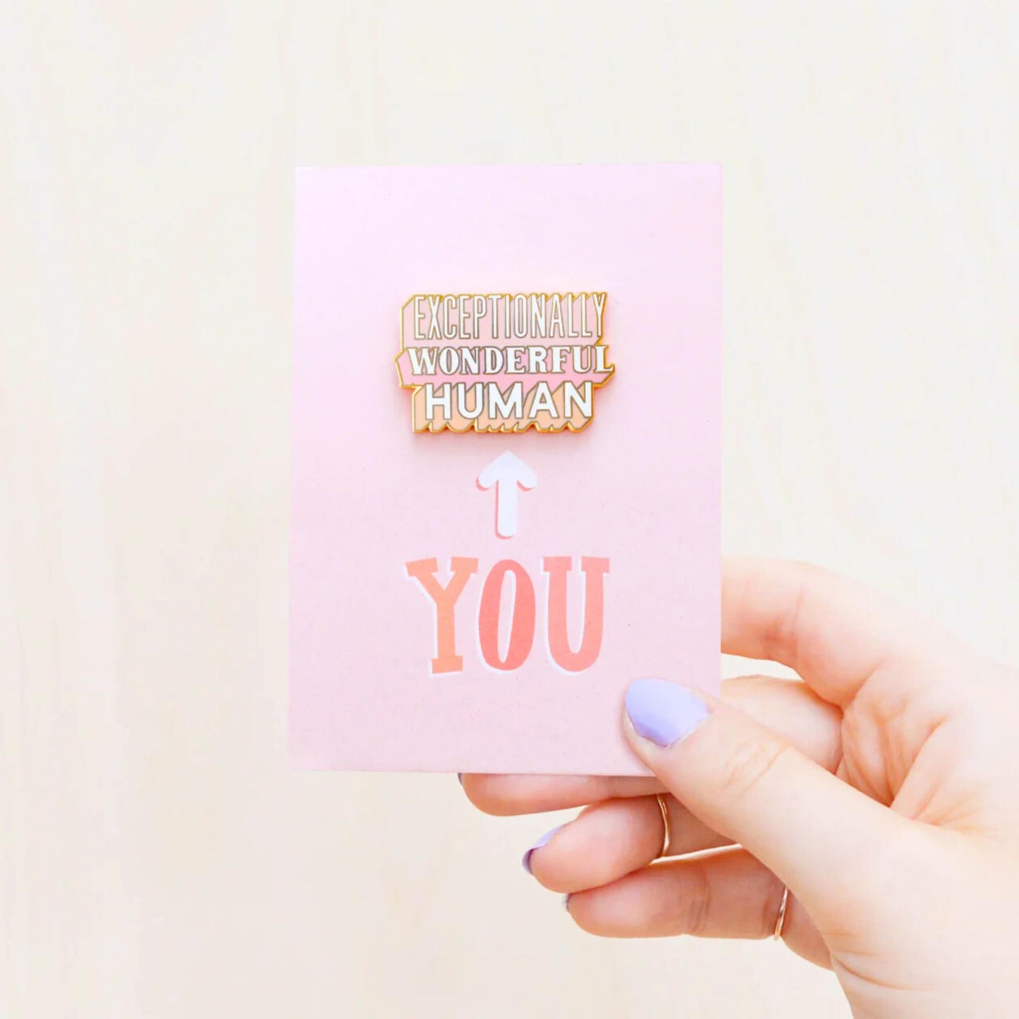 Hand holding Exceptionally Wonderful Human enamel pin badge on pink card.