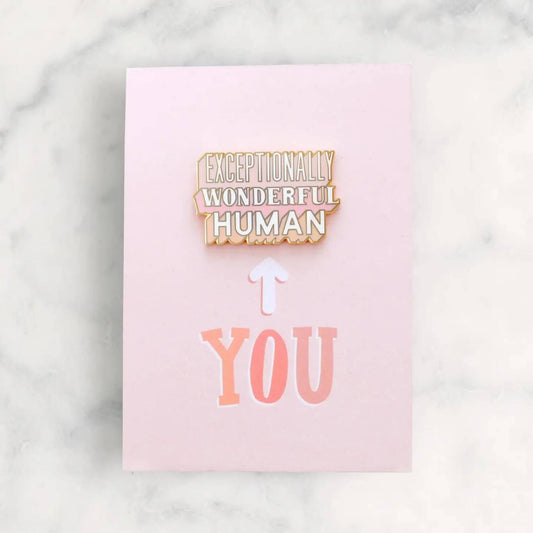 Exceptionally Wonderful Human enamel pin badge displayed on pink recycled backing card against marble surface.