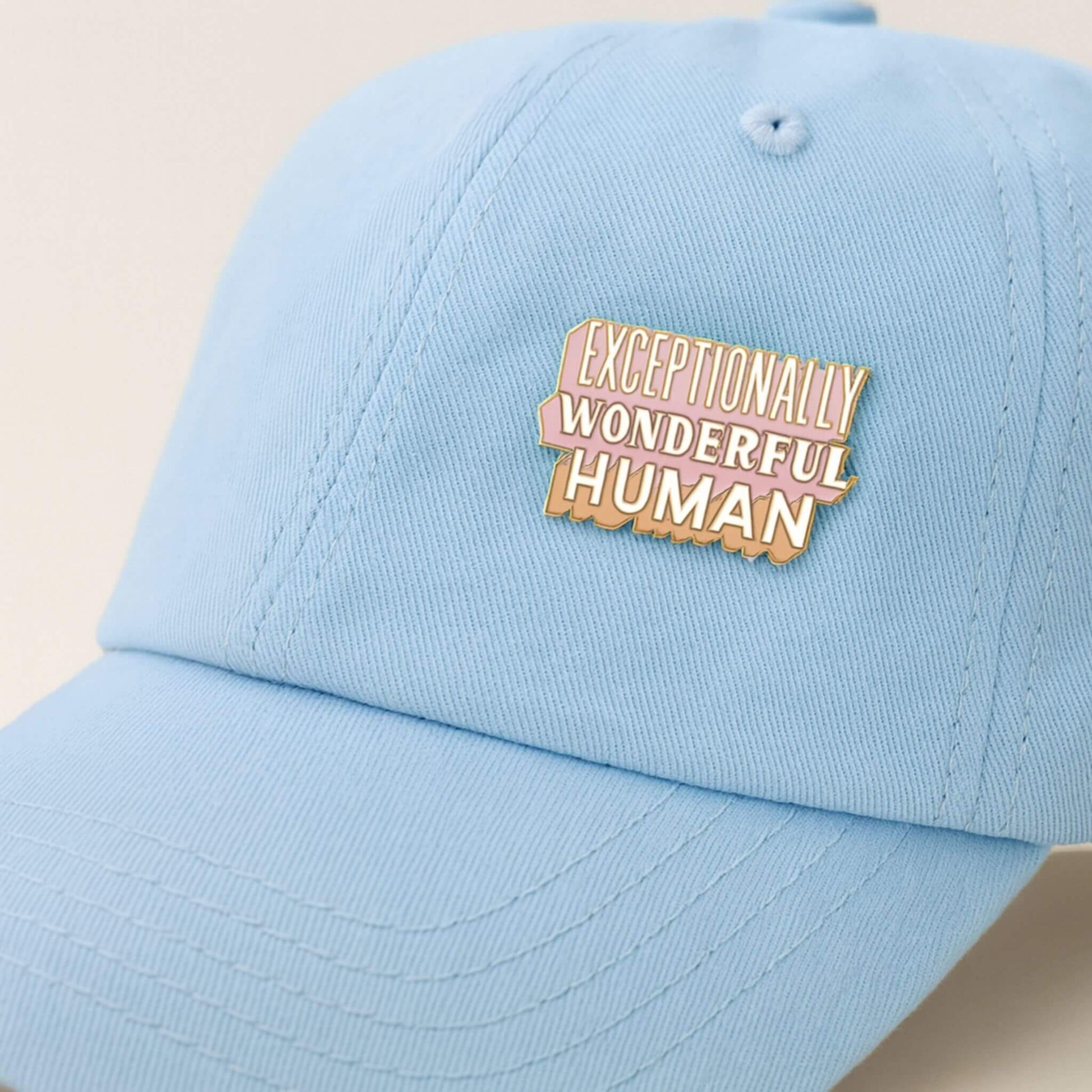 Light blue cap with 'Exceptionally Wonderful Human' badge on a beige background