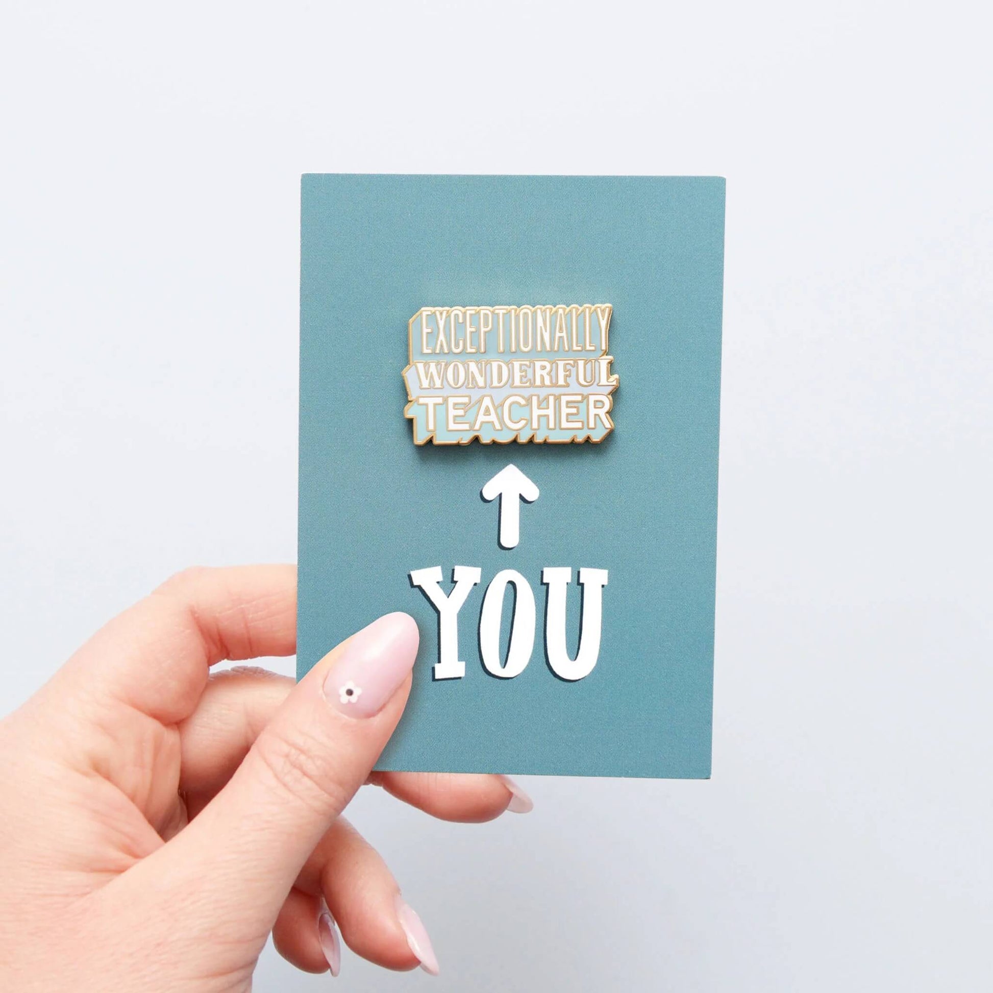 Hand holding Exceptionally Wonderful Teacher enamel pin badge on blue recycled backing card.