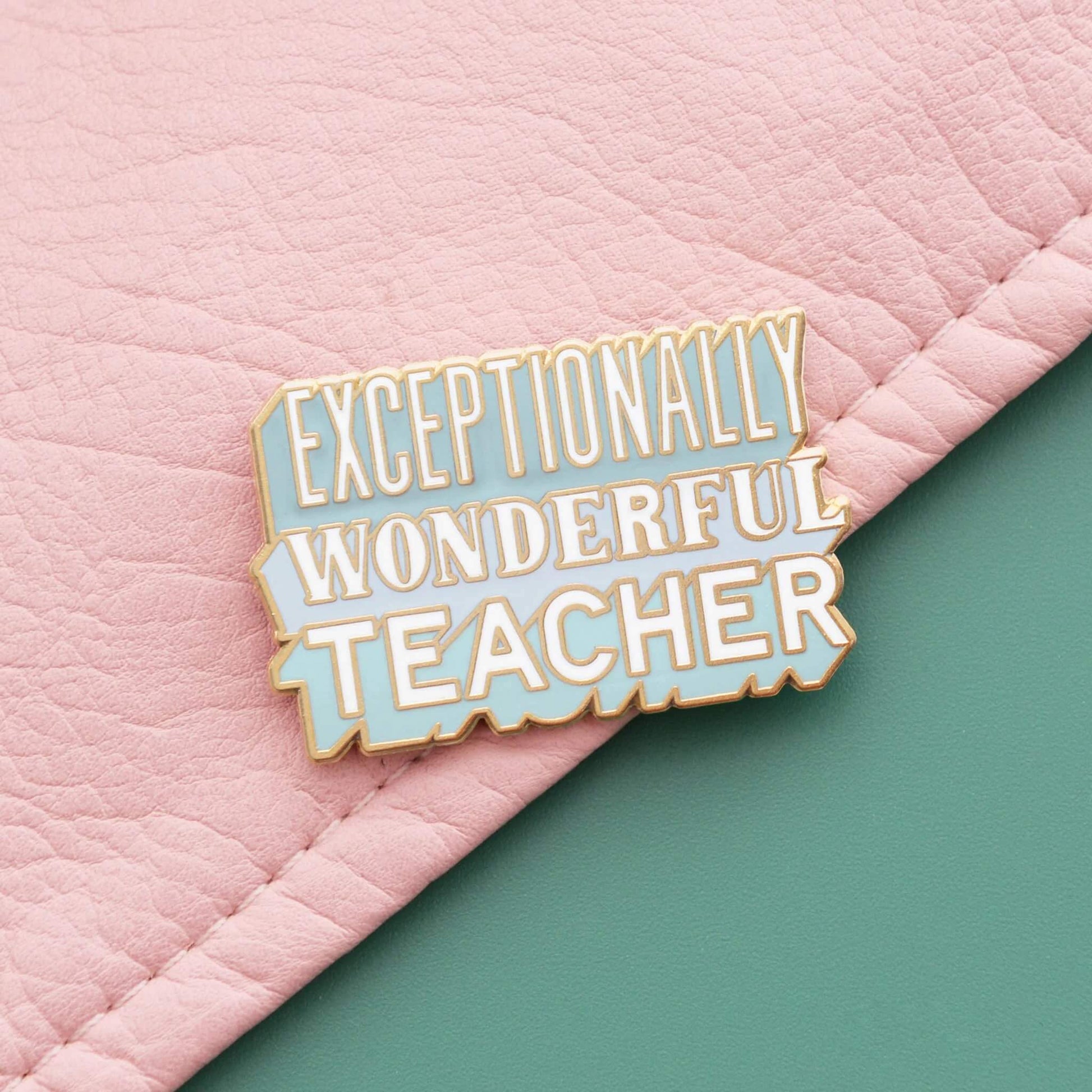 Exceptionally Wonderful Teacher enamel pin badge with matt gold finish placed on pink textured fabric and a green background.