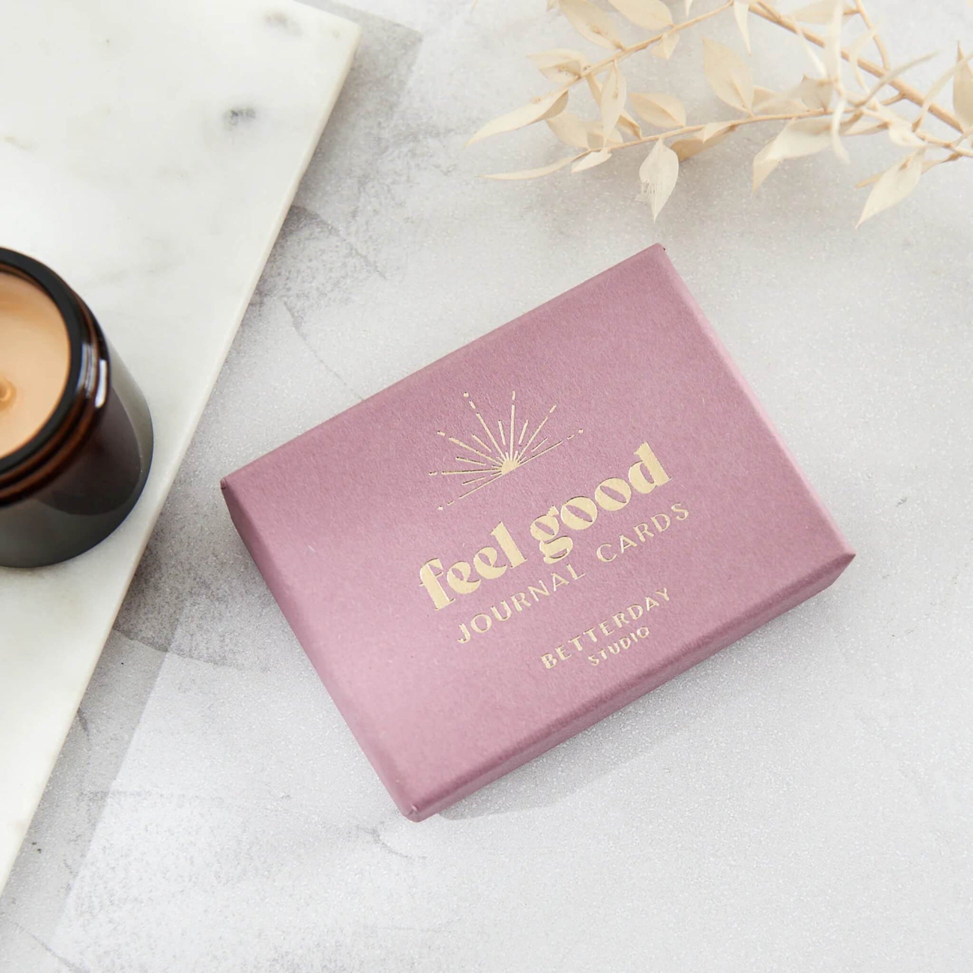 Pink box with gold lettering that says "Feel Good Journal Cards' on a white background with a lit candle
