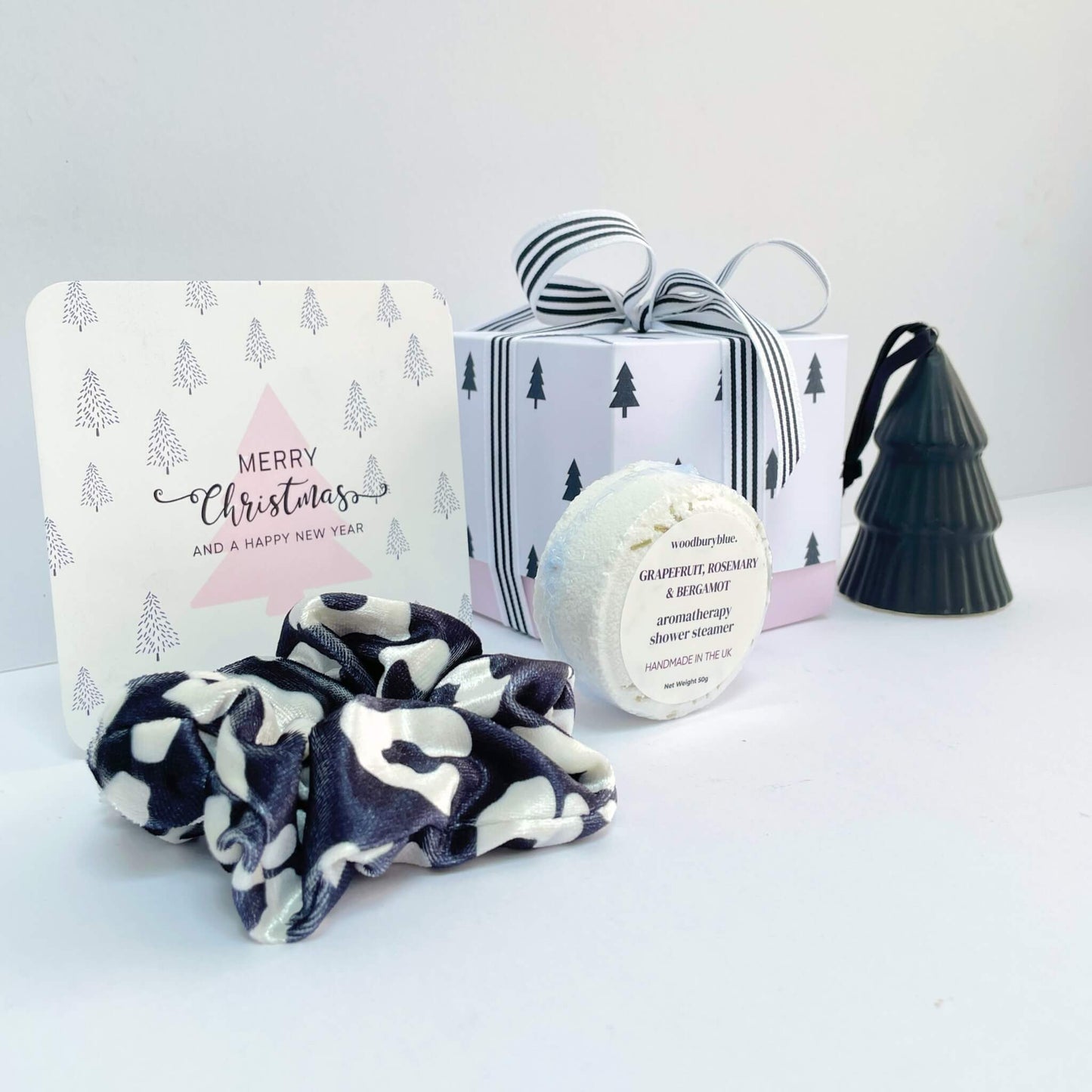 Angled view of the Little Box of Festive Treats featuring a grapefruit rosemary shower steamer, monochrome scrunchie, pink tree Christmas card, black tree ornament, and striped ribbon gift box.