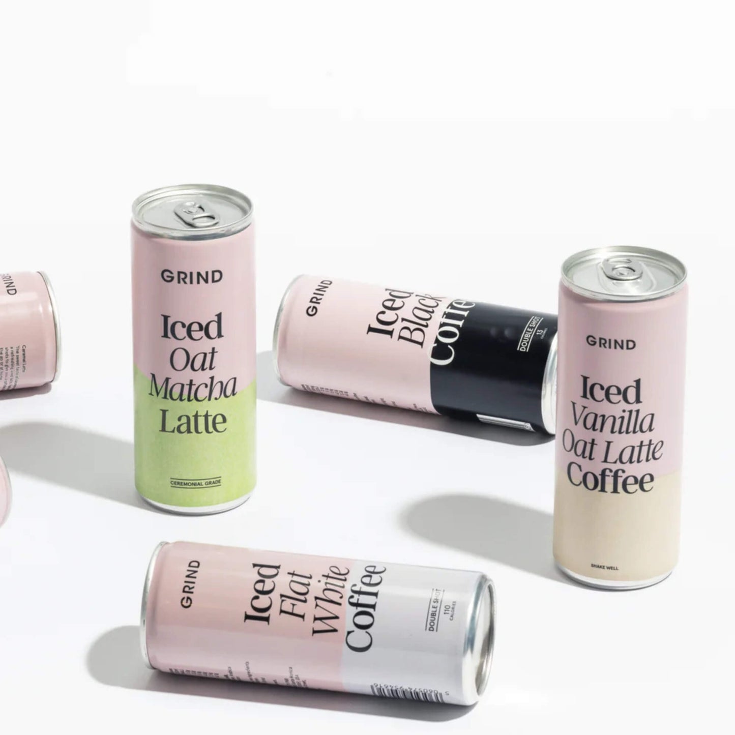 Lifestyle image showing several Grind iced coffee cans, including the Iced Vanilla Oat Latte alongside other flavours laid out on a white background.