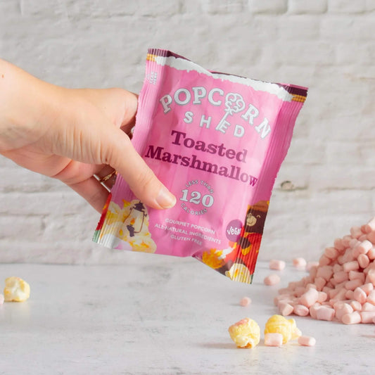 Hand holding vegan Toasted Marshmallow popcorn snack pack with scattered popcorn and marshmallow pieces.