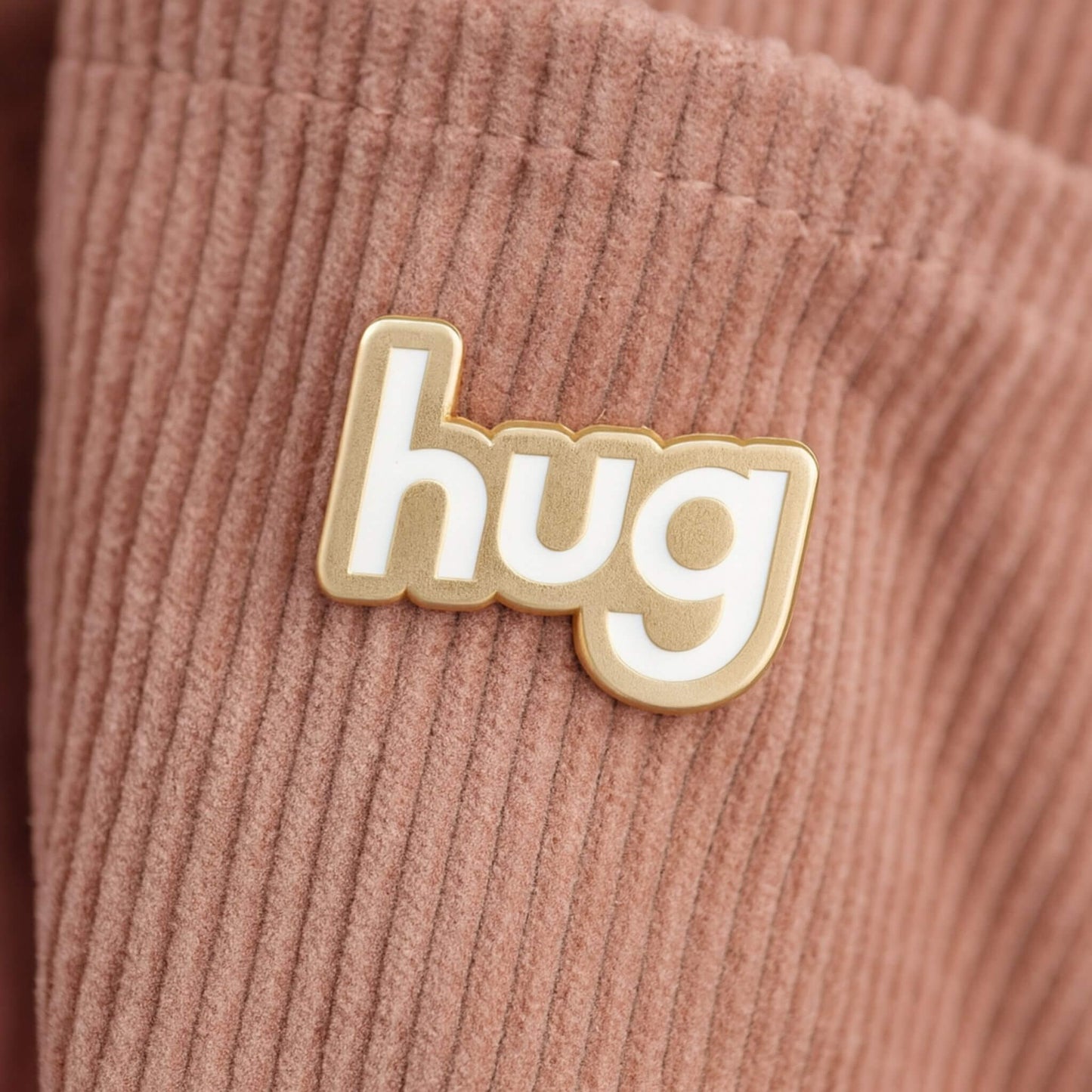 Hug enamel pin badge with gold outline and white lettering attached to a blush pink corduroy bag.