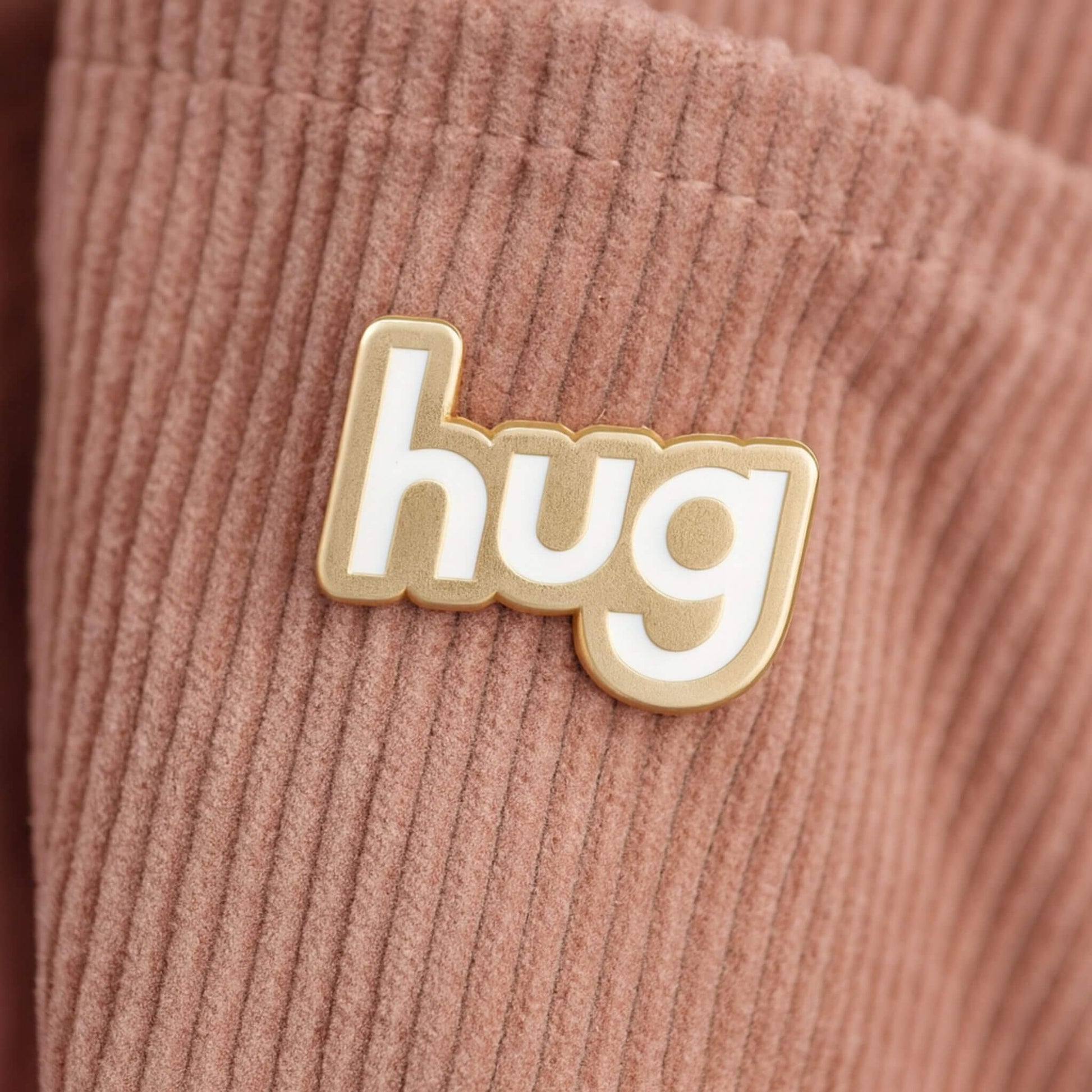 Hug enamel pin badge with gold outline and white lettering attached to a blush pink corduroy bag.