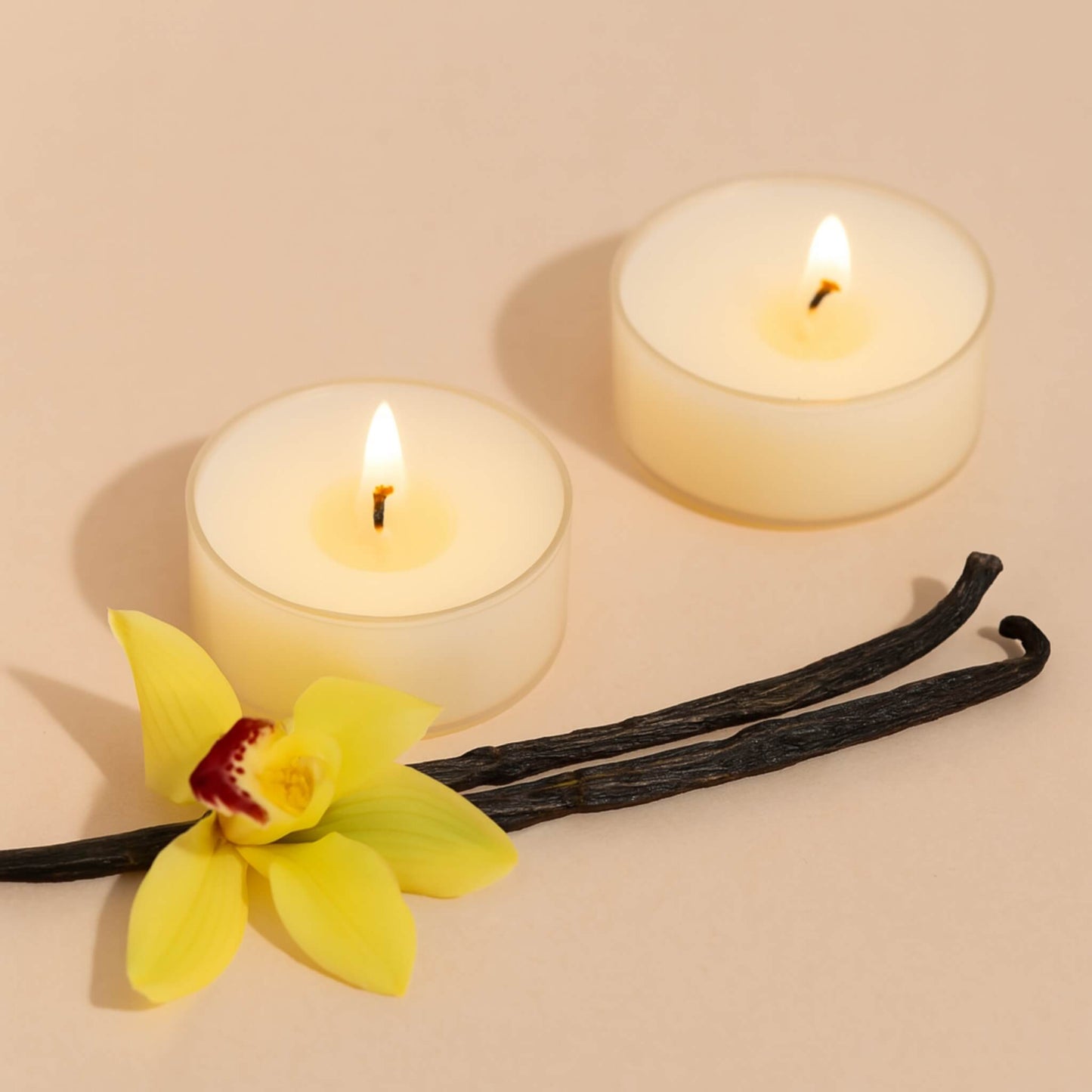 Two lit tea light candles, a vanilla bean, and a yellow flower on a beige background