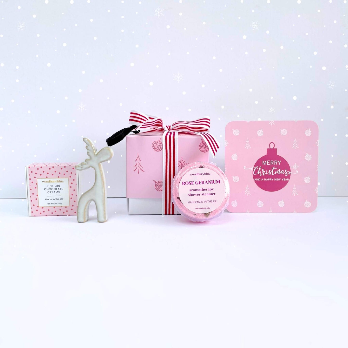 Merry & Bright Christmas Gift Box in pink with a striped ribbon, rose geranium shower steamer, ceramic reindeer ornament, pink gin chocolate creams, and a pink bauble card in a snowy scene