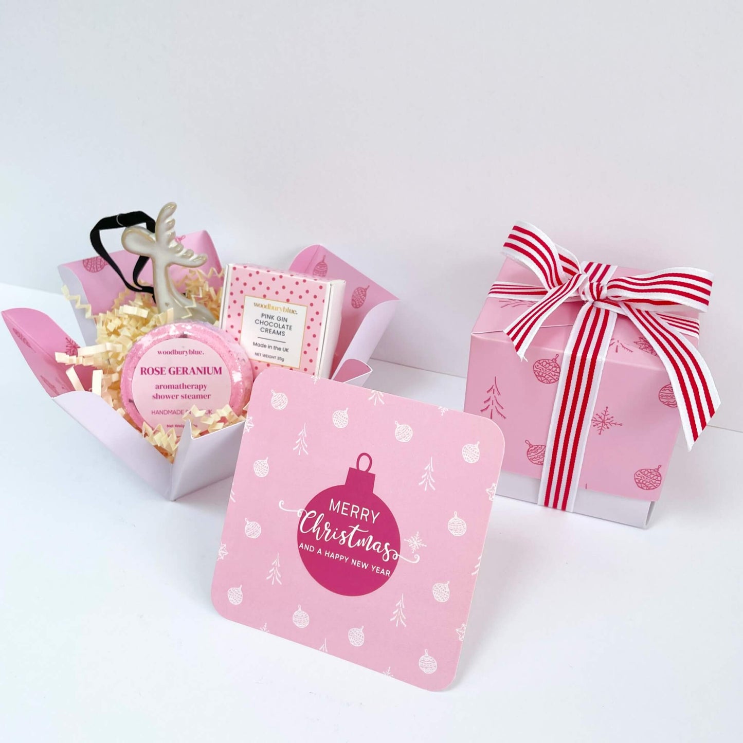 Unboxed Merry & Bright Gift Box featuring rose geranium shower steamer, ceramic reindeer ornament, pink gin chocolate creams, and a pink bauble Christmas card with the gift box and red striped ribbon in the background.