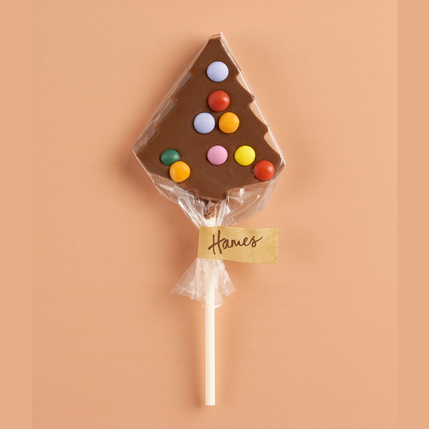Hames milk chocolate Christmas tree lollipop decorated with colourful candy beans, wrapped in clear cellophane with gold Hames label on a soft peach background