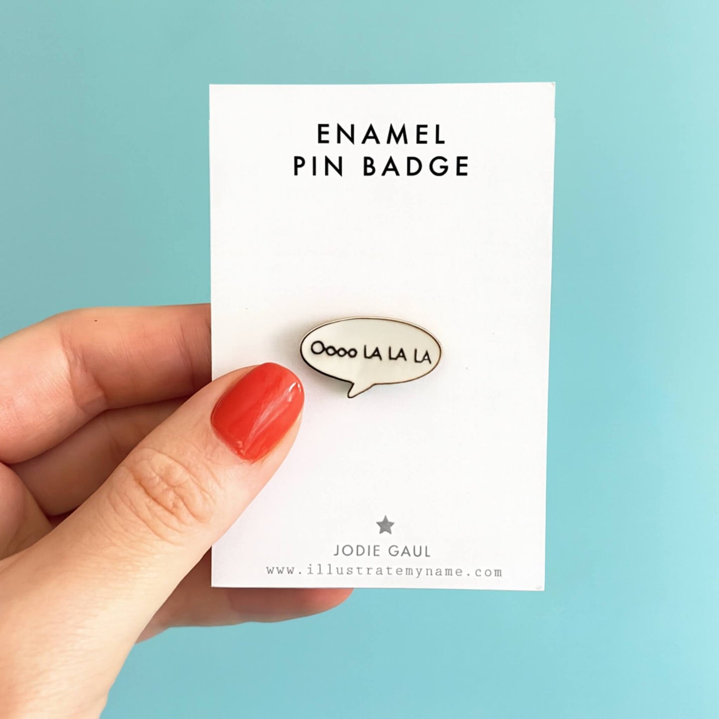 woman’s hand with red painted nails holding a white card displaying the Oooo La La La enamel pin badge against a pastel blue background.