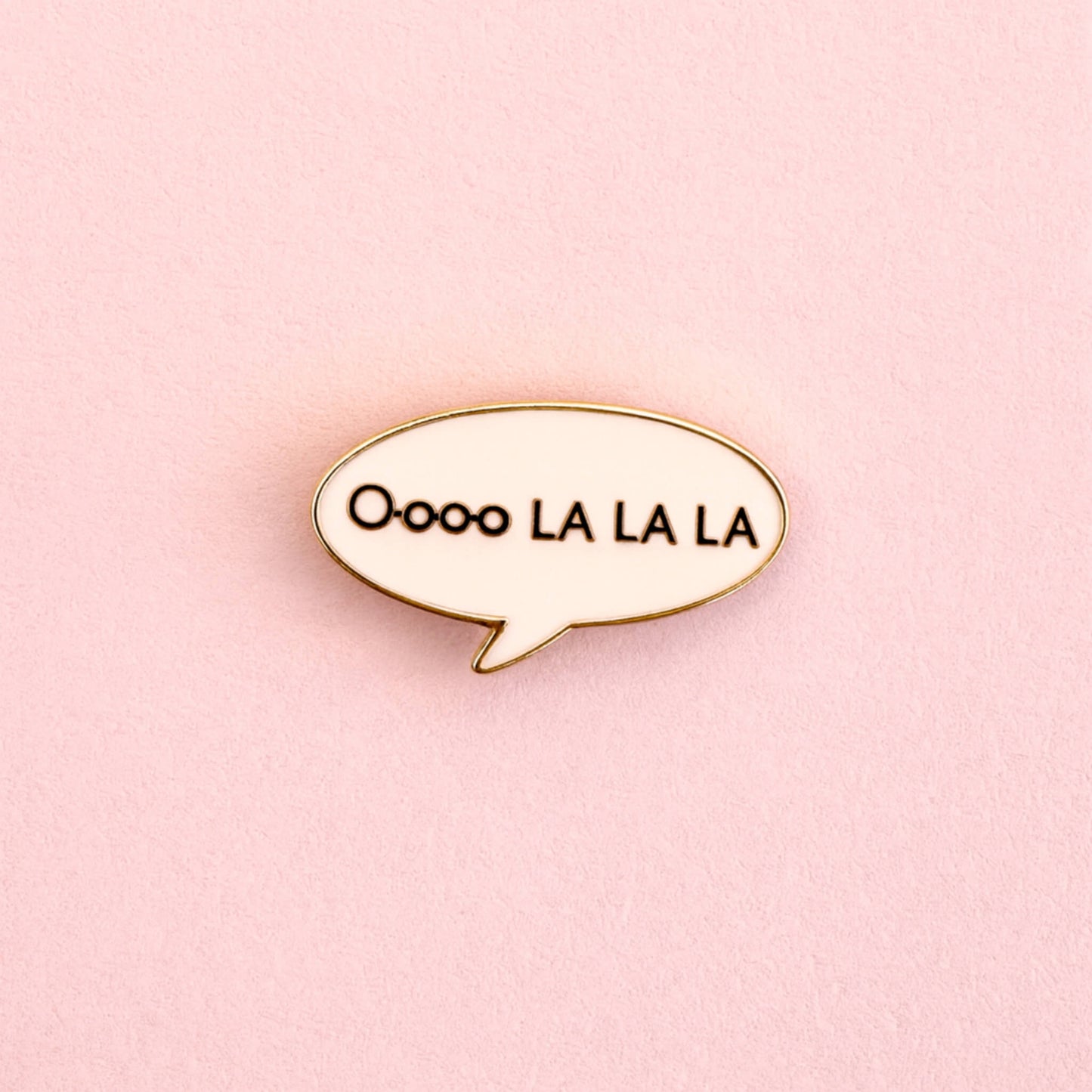 Oooo La La La enamel pin badge in a gold-outlined speech bubble, photographed on a pastel pink background.