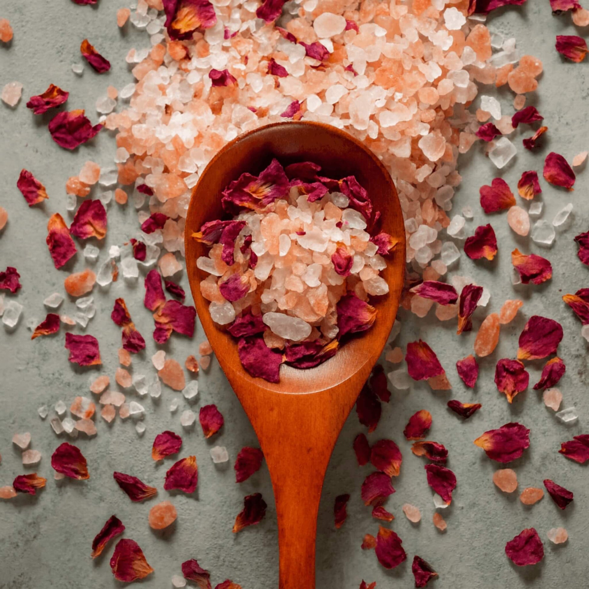 Restore aromatherapy bath salts with Himalayan pink salt and dried rose petals in a wooden spoon on grey surface