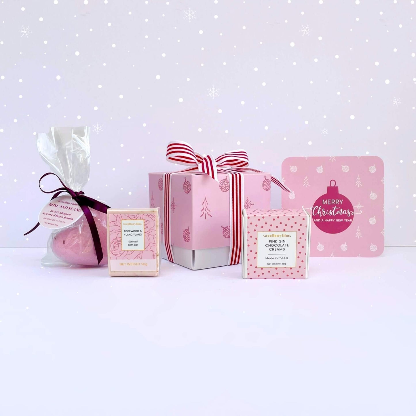 Rosy Glow Christmas Gift Box with pink bath bomb, rosewood soap, pink gin chocolate creams, pink ribbon gift box, and matching pink bauble Merry Christmas card on snowy background.