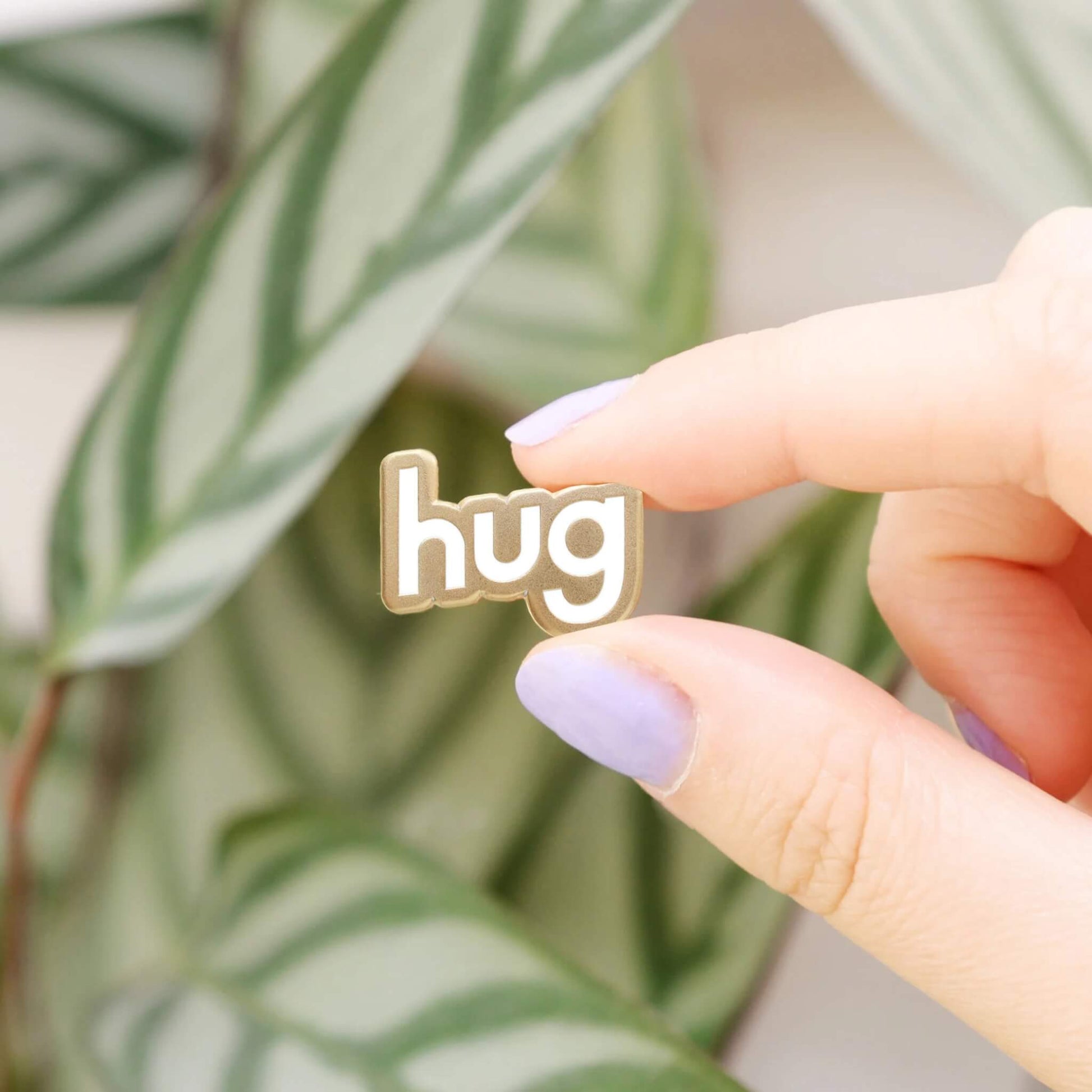 Close-up of Sending a Hug enamel pin badge held between fingers in front of green leaves.
