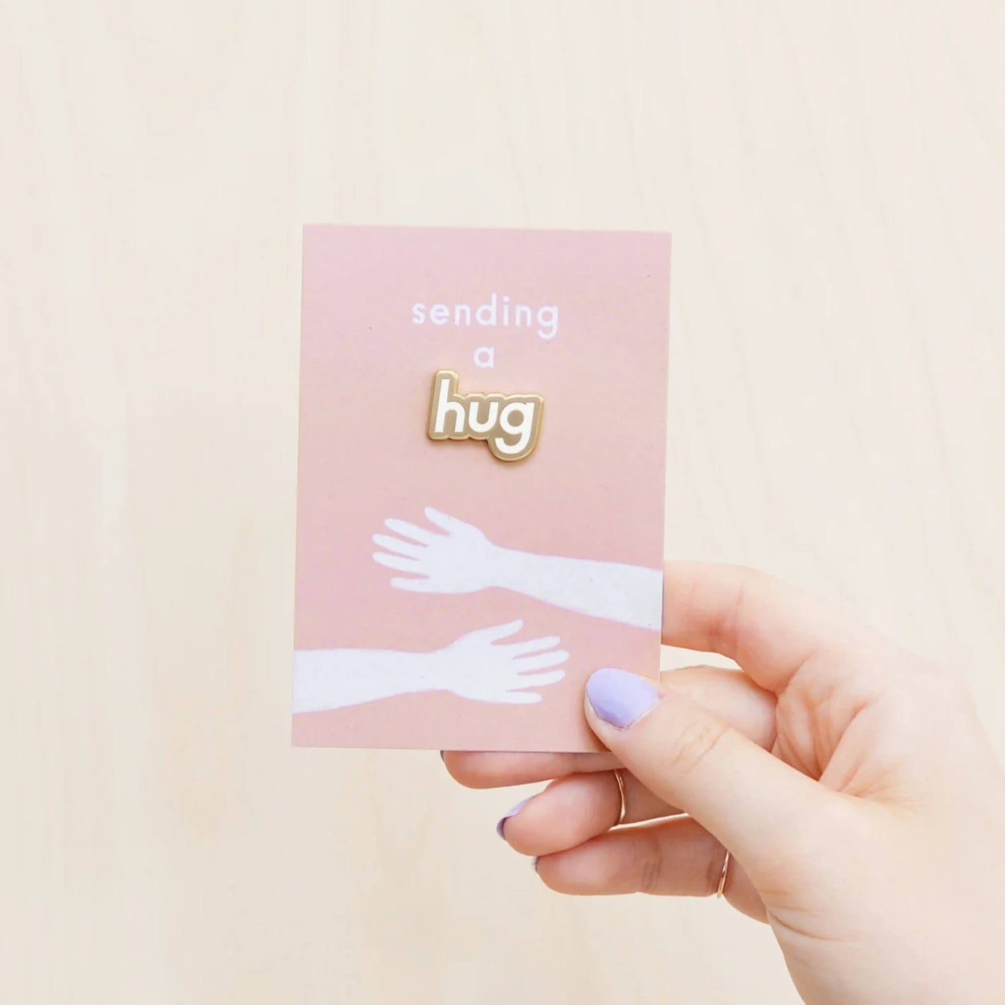 Hand holding Sending a Hug enamel pin badge on blush pink recycled backing card.