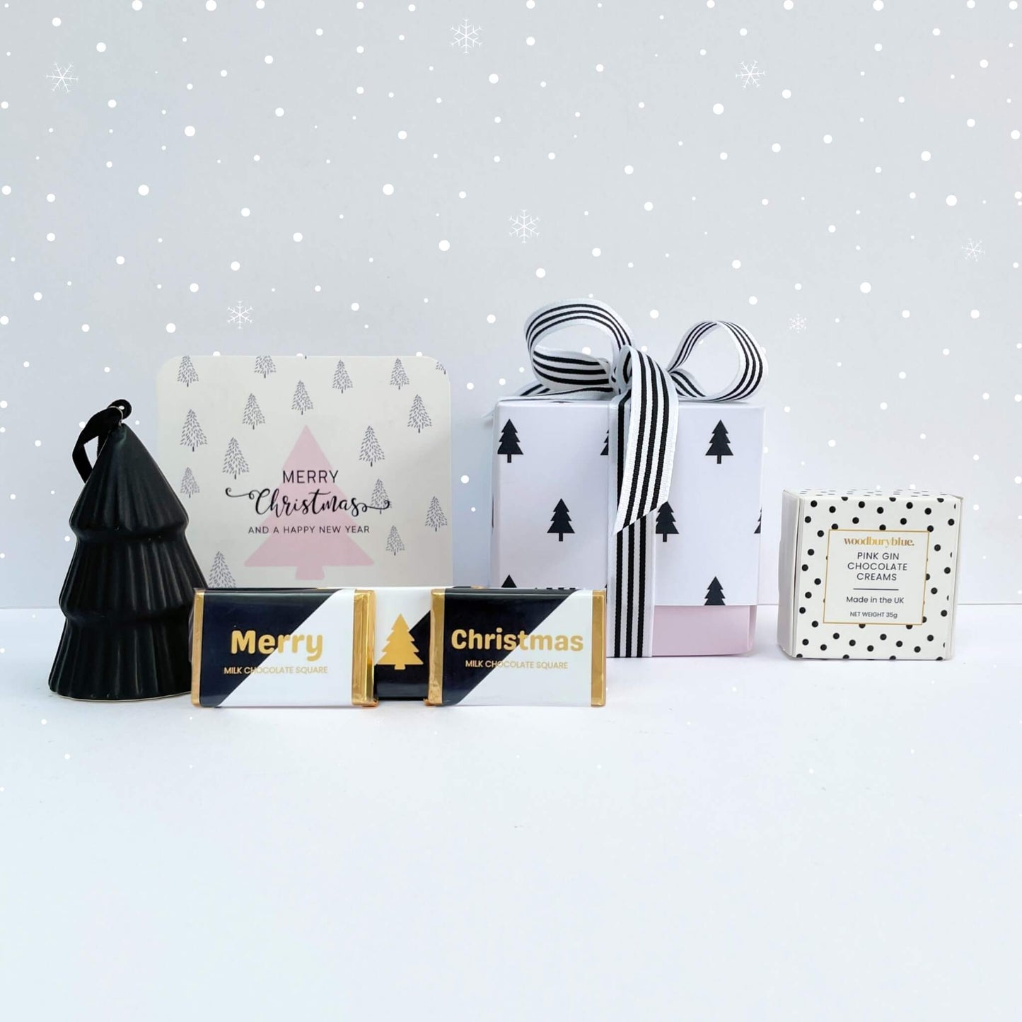White Christmas Gift Box styled with falling snow overlay, pink tree card, striped ribbon-wrapped gift box, black tree ornament, chocolate bars, and pink gin creams.