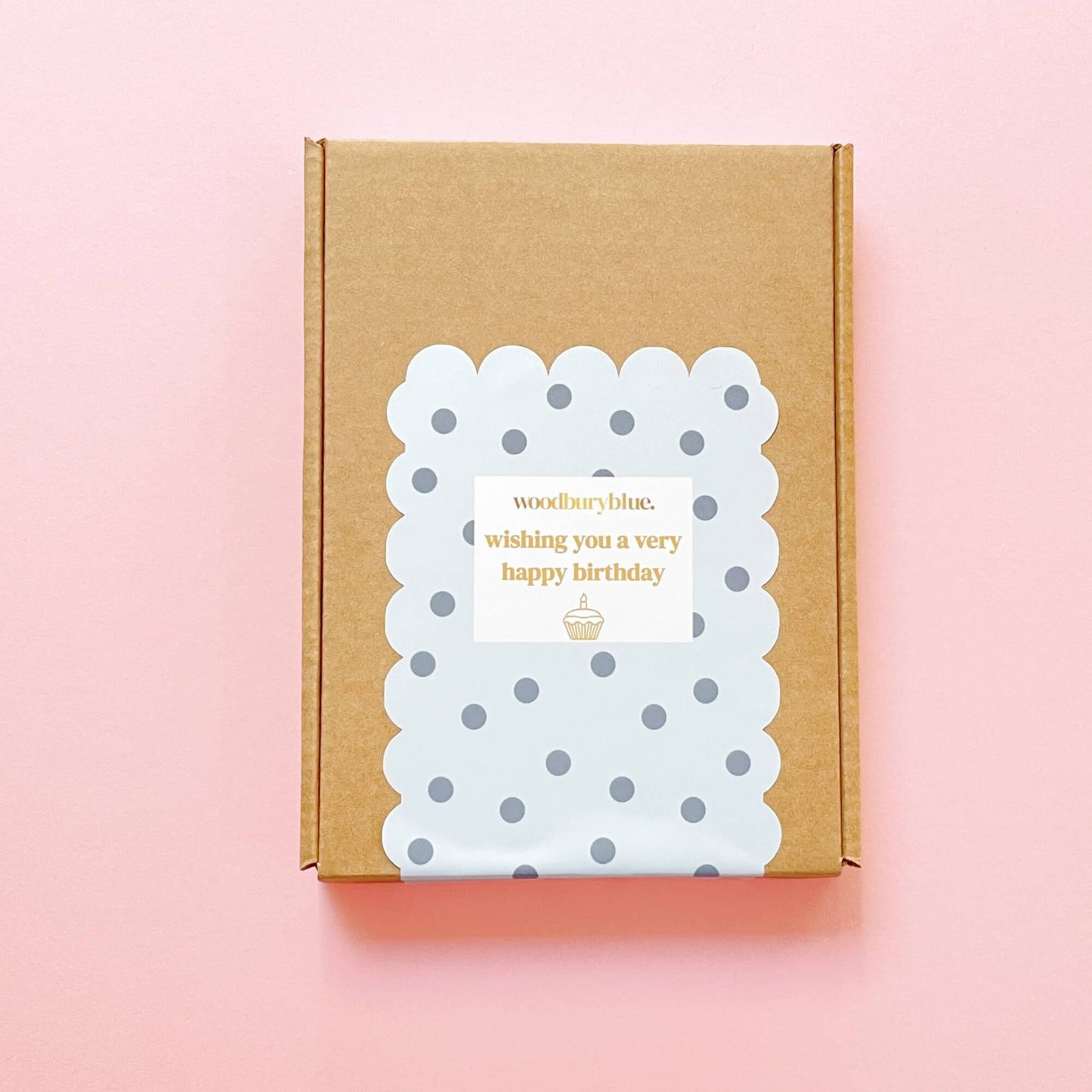 Closed Woodbury Blue Birthday Wishes Socks and Chocs Letterbox Gift box with grey polka dot scalloped sticker and gold text reading “Wishing you a very happy birthday” on a pink background.