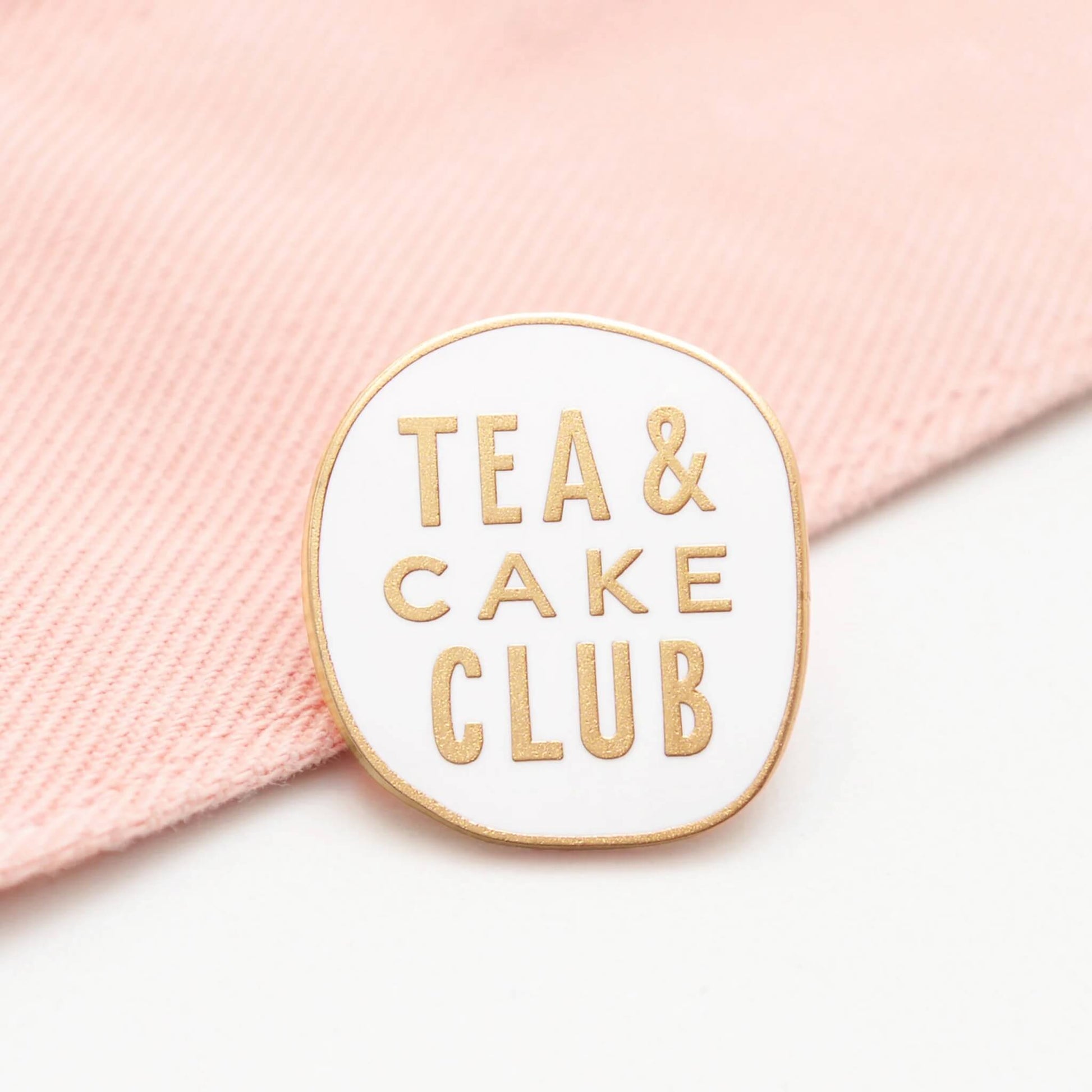 Tea and Cake Club enamel pin badge with gold lettering on white enamel, resting on pale pink fabric background