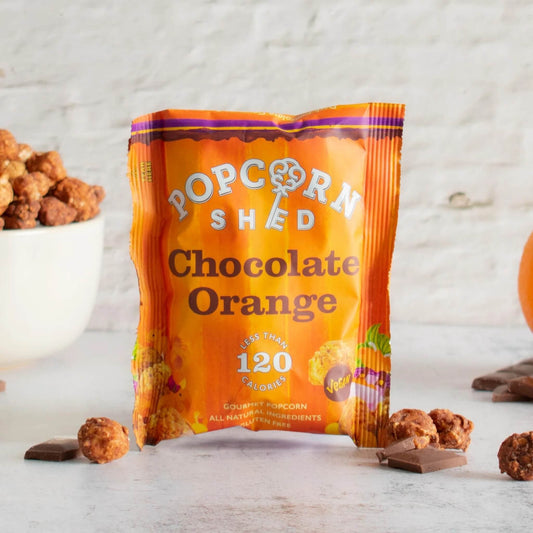 Popcorn Shed Vegan Chocolate Orange Gourmet Popcorn 24g sachet standing on light background with dark chocolate and orange slices nearby.