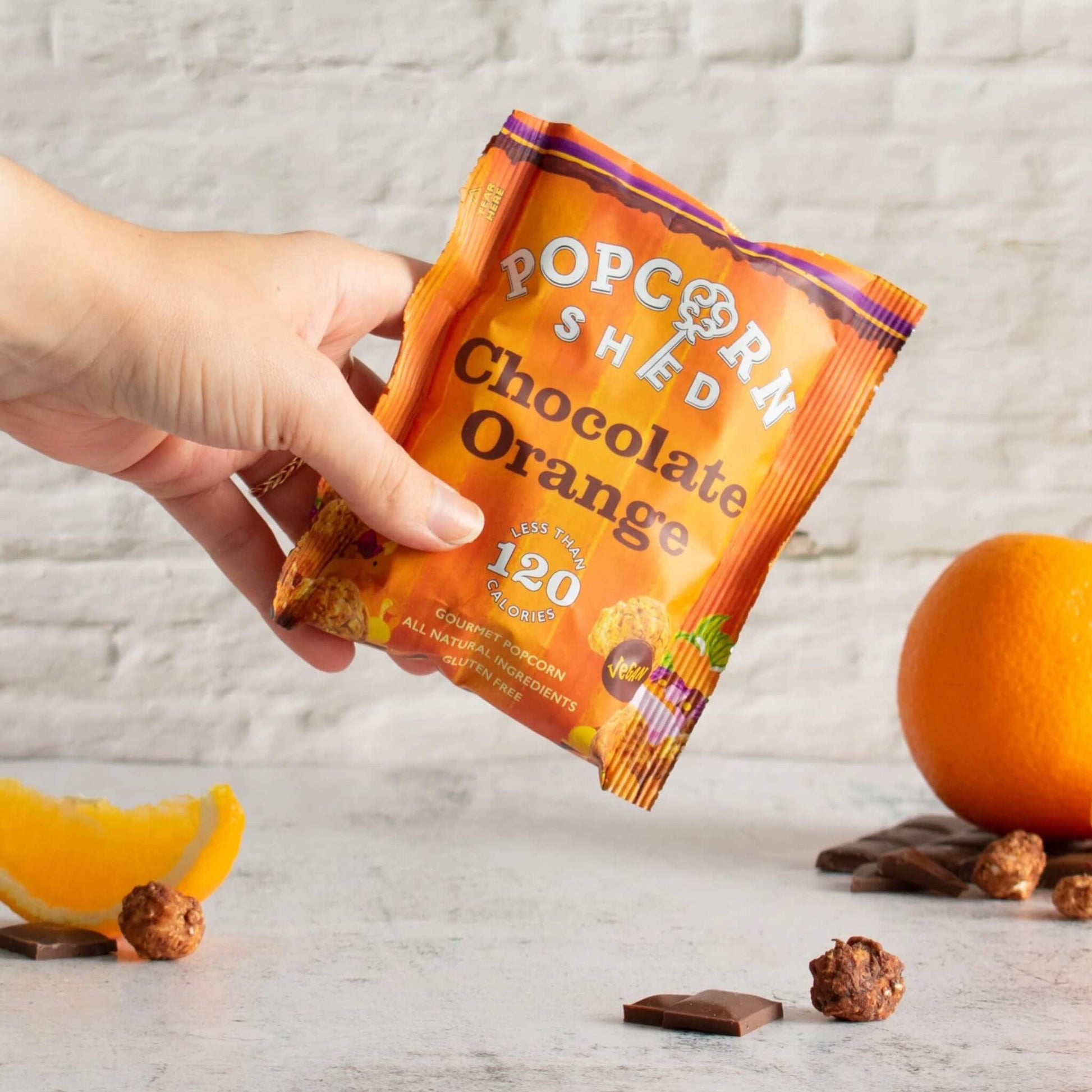 Hand holding Popcorn Shed Vegan Chocolate Orange Gourmet Popcorn pack with pieces of popcorn, chocolate, and orange in the background.