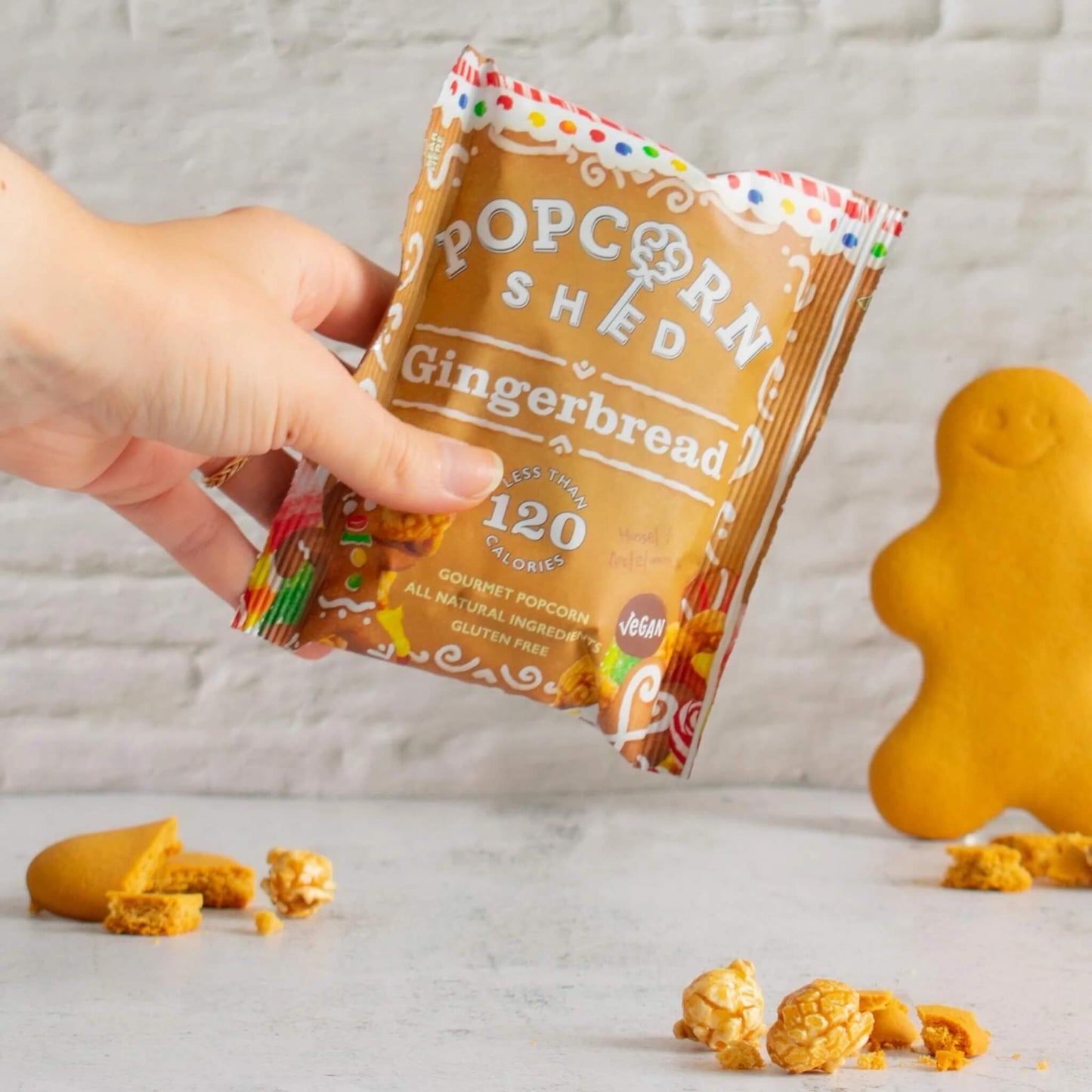 Hand holding Popcorn Shed Vegan Gingerbread Gourmet Popcorn snack pack with gingerbread biscuits in the background.