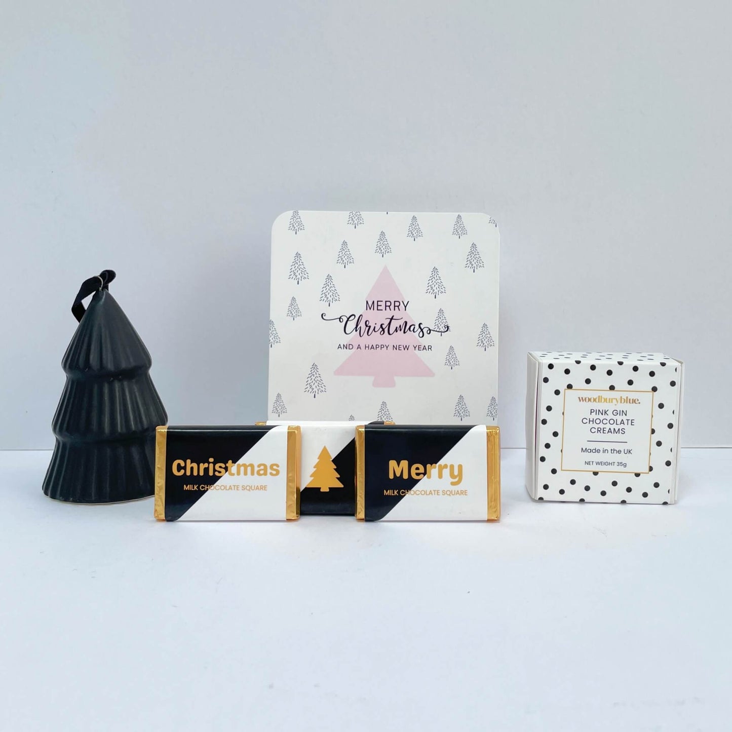 Close-up of the White Christmas Gift Box arrangement featuring a black tree ornament, Merry Christmas card, two mini chocolate squares, and a box of pink gin chocolate creams.