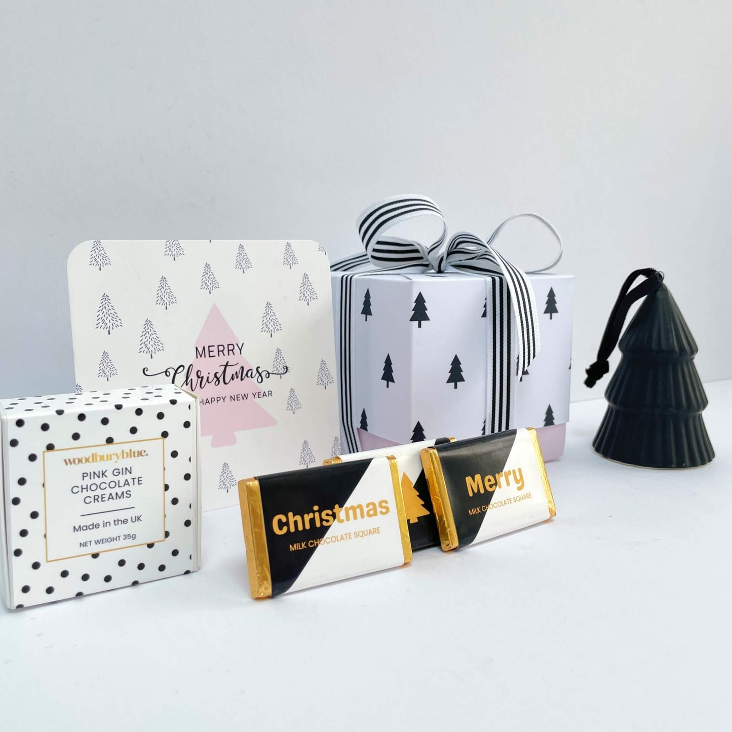 Side view of the White Christmas Gift Box with black tree gift wrap, striped ribbon, pink tree Christmas card, two chocolate squares, a box of pink gin chocolate creams, and a black ceramic tree ornament.