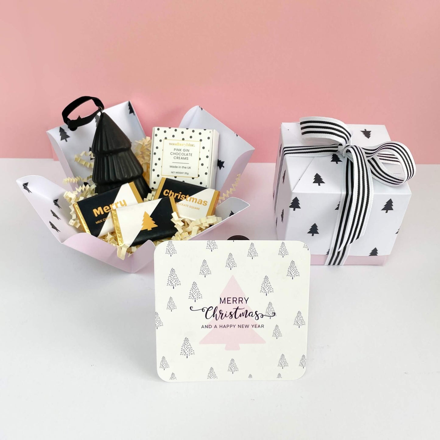 Unboxed White Christmas Gift Box with three mini milk chocolate squares, pink gin chocolate creams, black ceramic Christmas tree ornament, and pink tree Christmas card beside the closed ribbon-tied box.