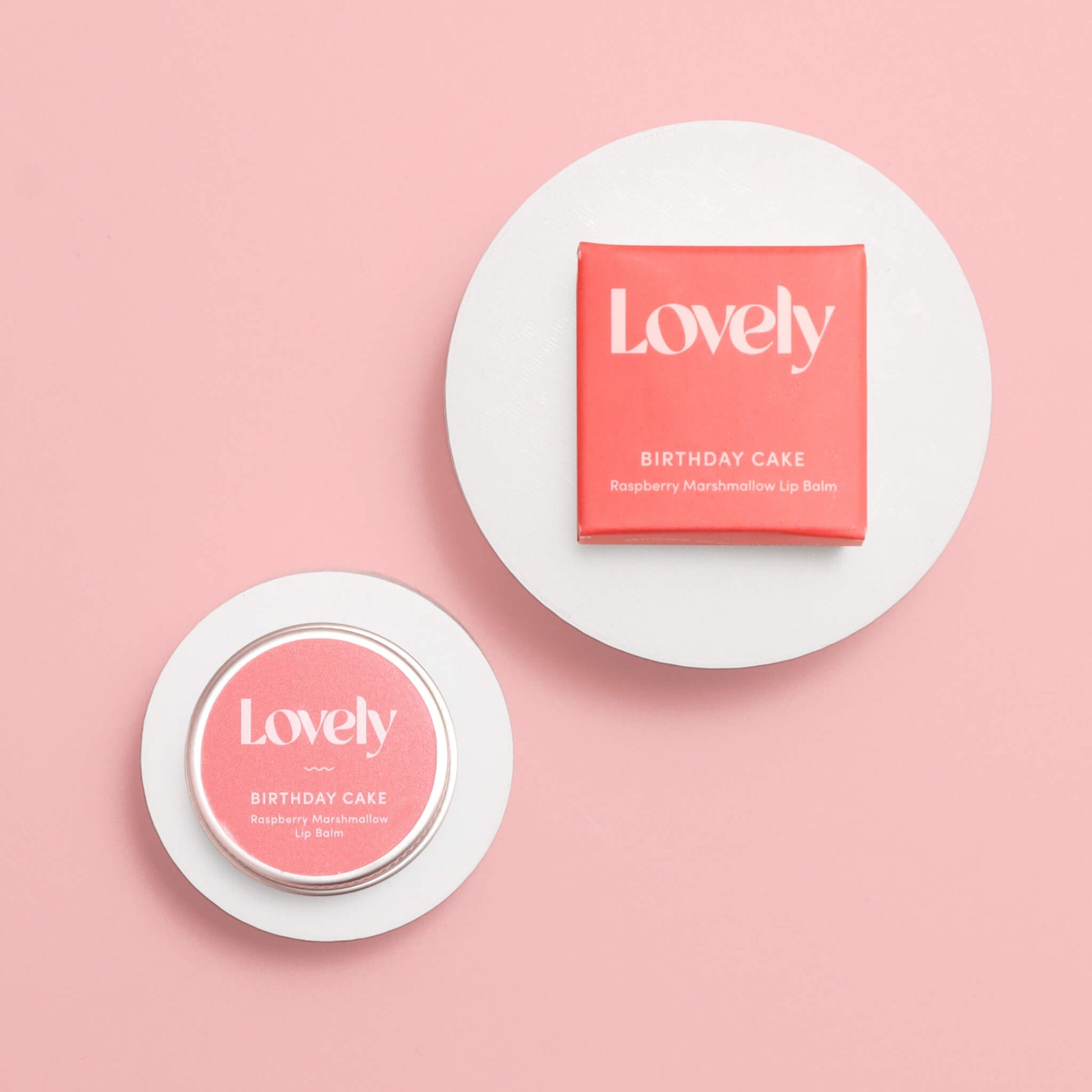 Lovely Skincare Birthday Cake Raspberry Marshmallow Lip Balm tin and box on circular white stands against pink background