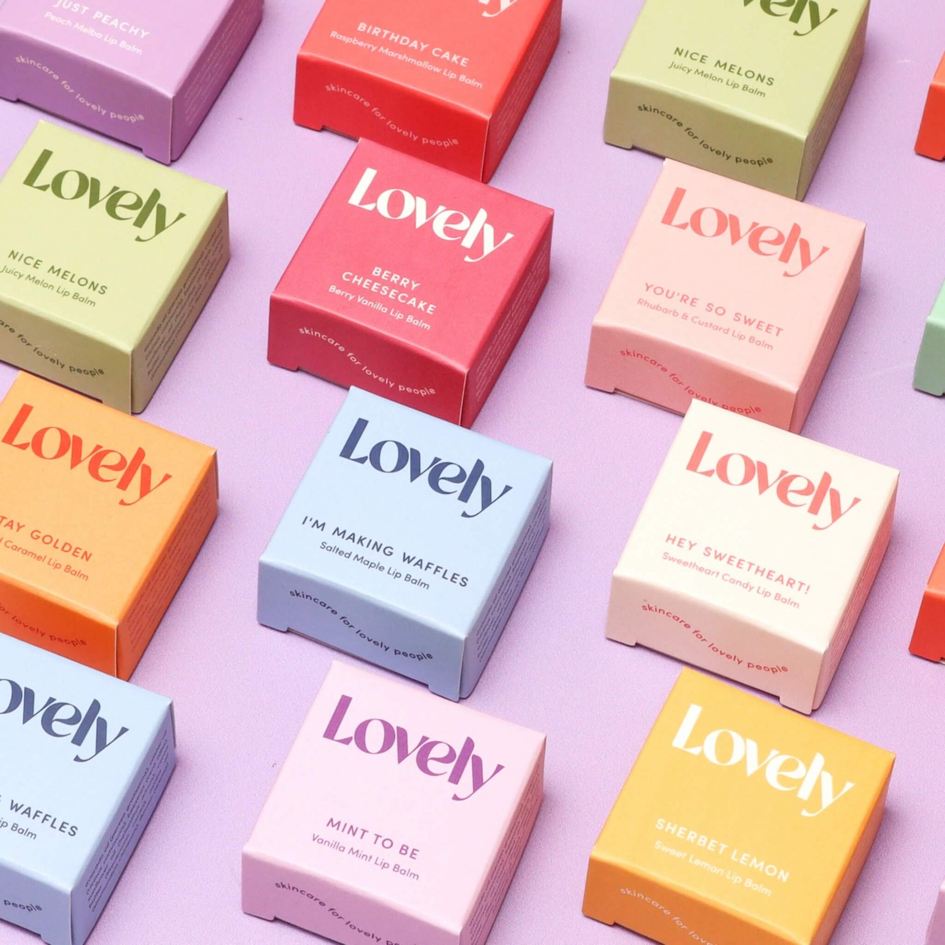Lovely Skincare Sherbet Lemon Lip Balm among colourful lip balm boxes displayed on pastel purple background