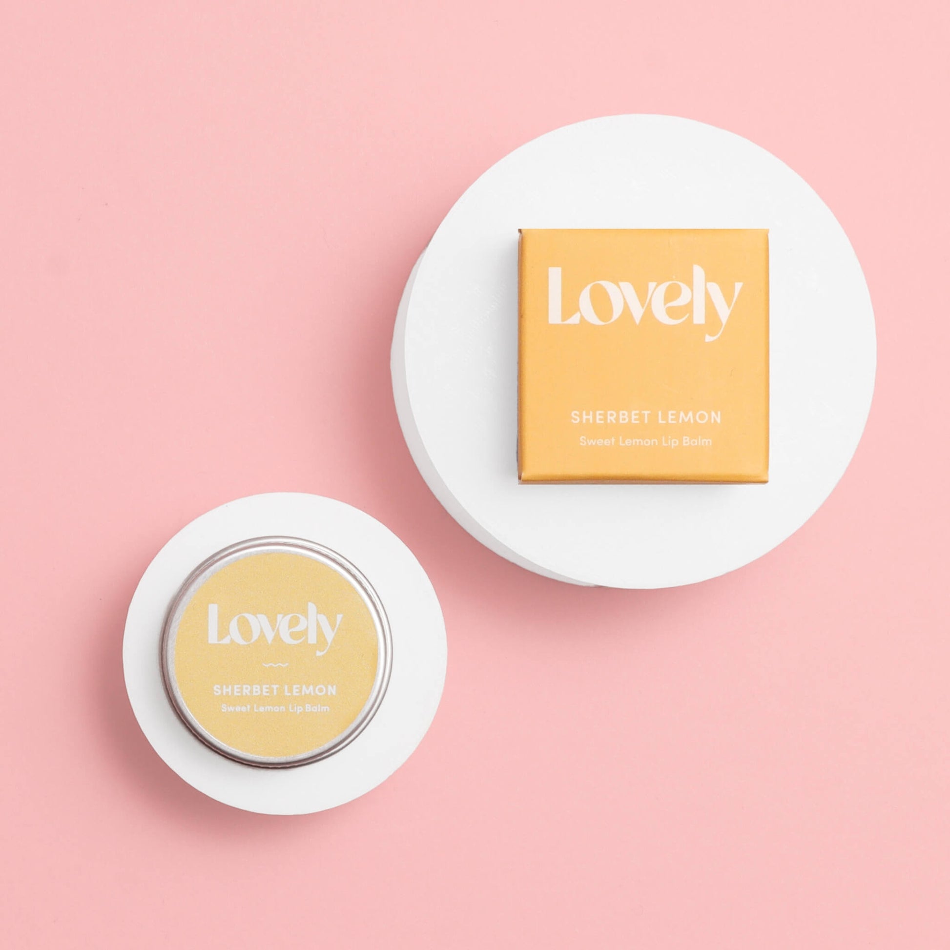 Lovely Skincare Sherbet Lemon Sweet Lemon Lip Balm tin and box displayed on circular white stands against pink background