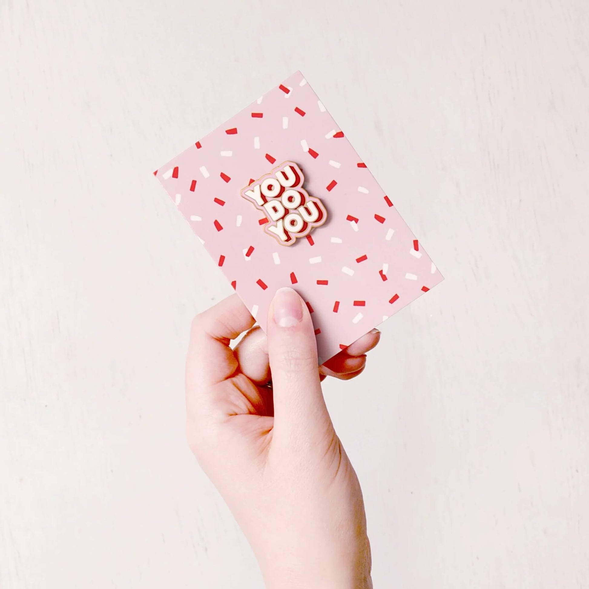 Hand holding a You Do You enamel pin badge with gold edge attached to a pink backing card with red and white confetti pattern
