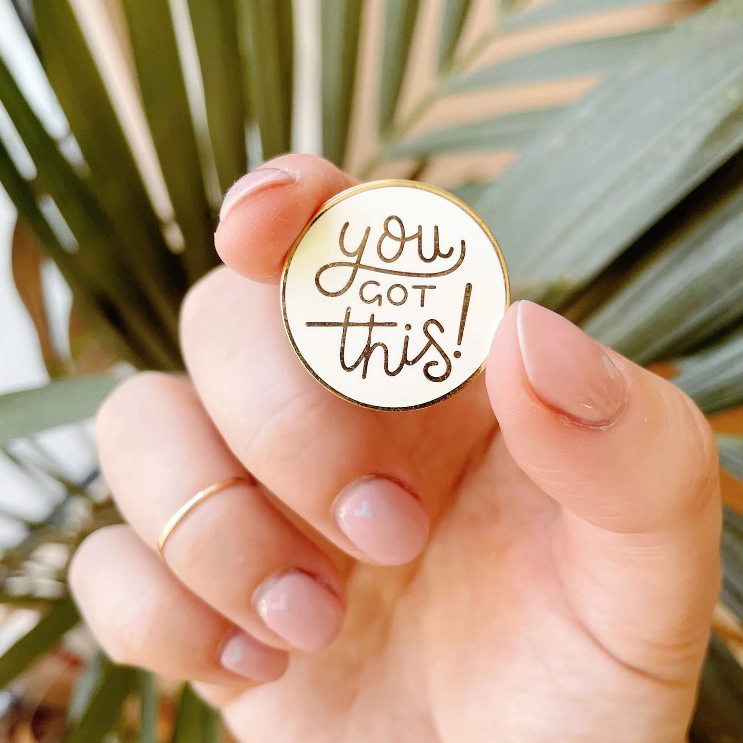 Close-up of You Got This enamel pin badge held between fingers in front of green leaves.