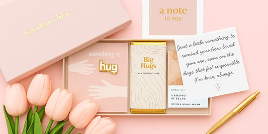 Flatlay of Woodbury Blue cheer up gift box with chocolate, bath bar, enamel pin and handwritten note on pink background with tulips – thoughtful letterbox gift for a friend