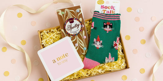 Socks and chocolate letterbox gift from Woodbury Blue, featuring a Spotty chocolate bar, green cat-print socks and a notecard presented in a kraft gift box with yellow shred on a pastel confetti background.