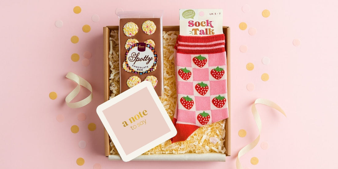 Socks and chocolate letterbox gift with strawberry socks, milk chocolate bar and a pink notecard, styled in a Kraft box with ribbon and confetti on a pale pink background