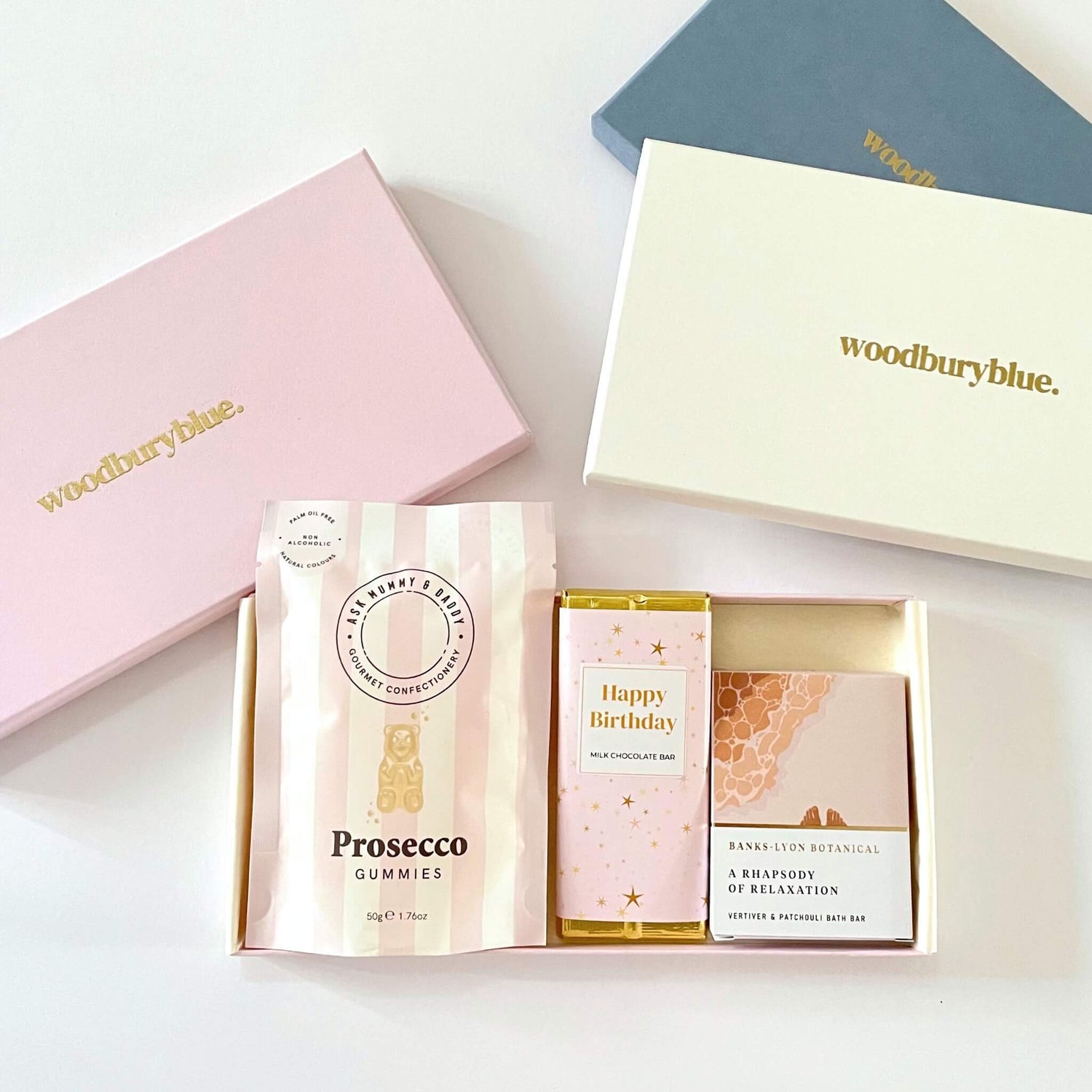 Letterbox gift set with Prosecco gummies, a Happy Birthday milk chocolate bar, and a botanical bath melt, presented in a cream Woodbury Blue gift box with pink and navy lids in background.