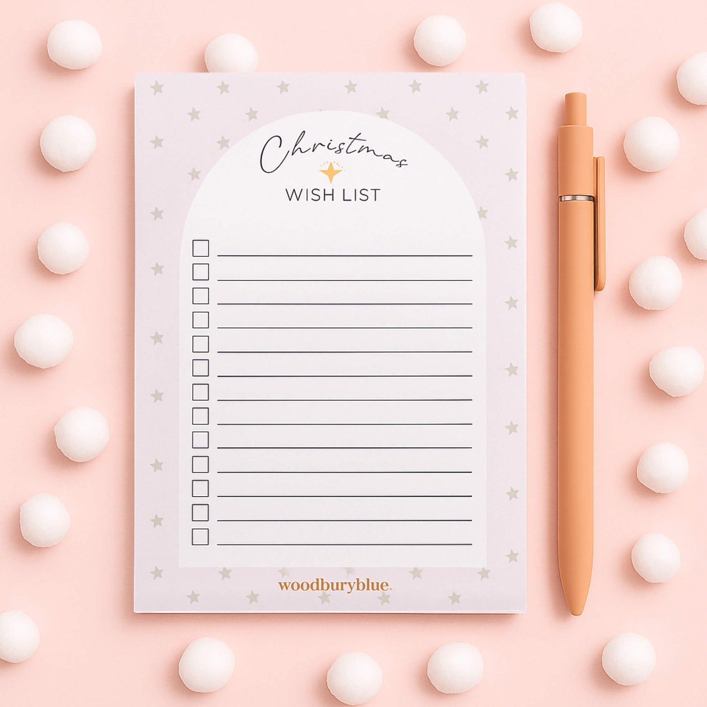 A6 Christmas Wish List Notepad by Woodbury Blue on a pale pink snowy background with fluffy white snowballs and a matching peach pen.